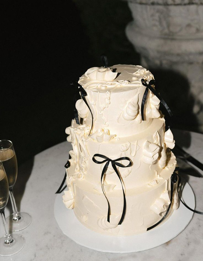 7 Wedding Cake Trends for 2025: Inspiring Designs for Modern Couples 13 White three-tier wedding cake with small black bows, photographed with flash.