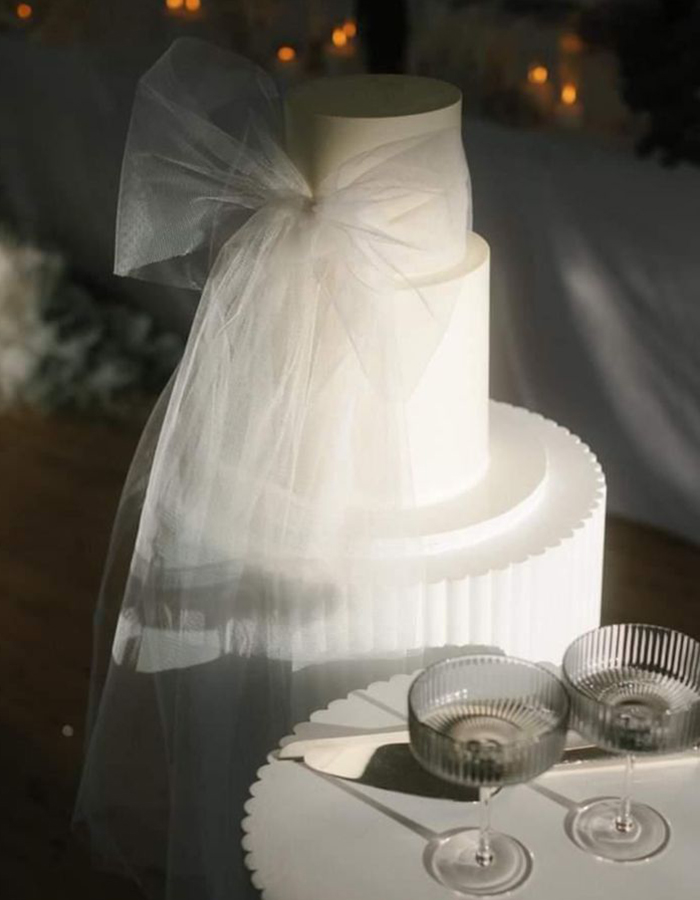 7 Wedding Cake Trends for 2025: Inspiring Designs for Modern Couples 12 White two-tier wedding cake with a giant white bow, photographed on a white cake stand.