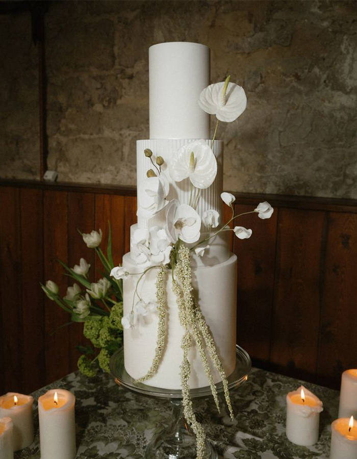 7 Wedding Cake Trends for 2025: Inspiring Designs for Modern Couples 6 Four-tier wedding cake with white fondant and floral details, photographed on a silver plate surrounded by candles.