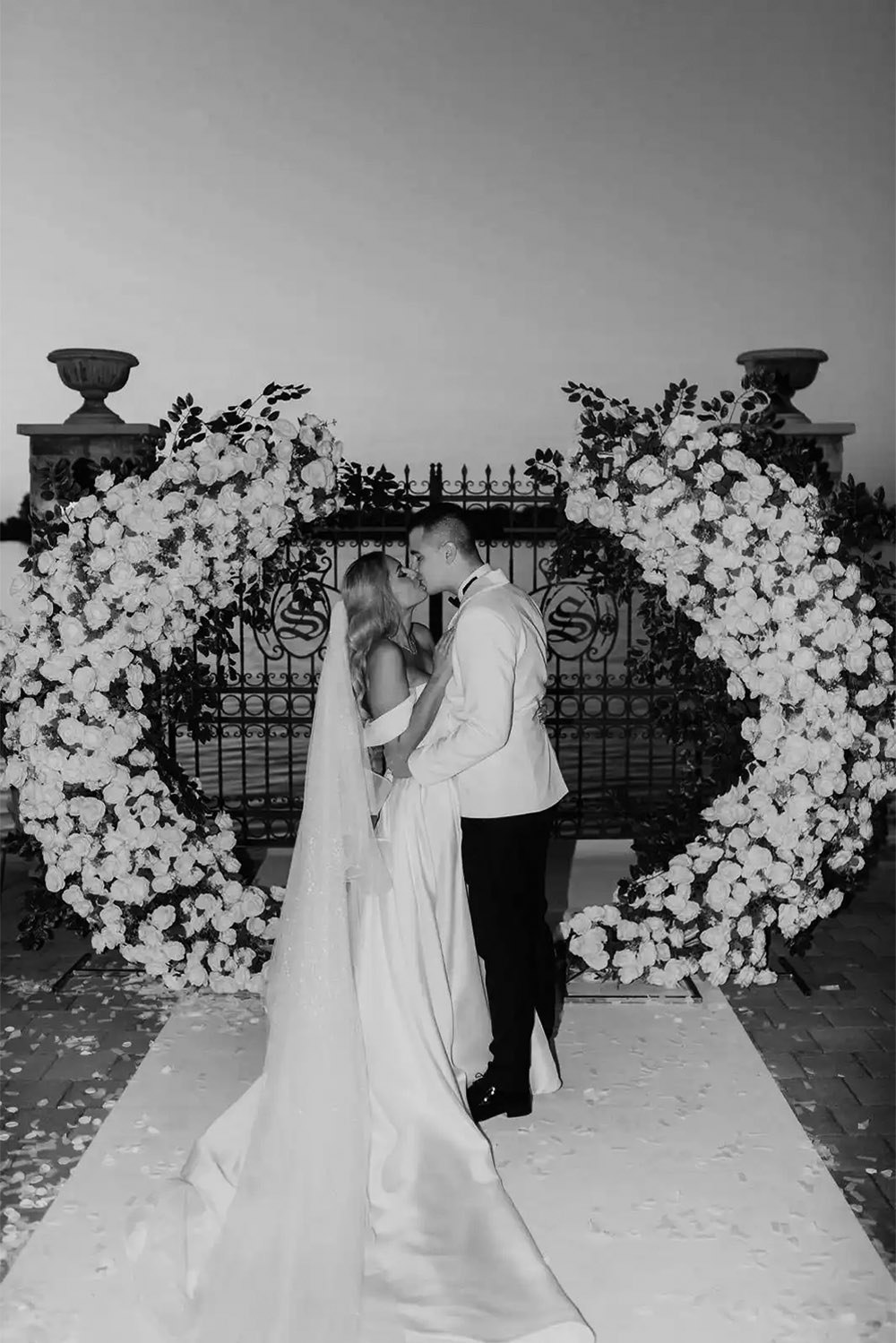 At a riverside wedding venue in Serbia, a newlywed couple shares a kiss beneath a dramatic floral installation, captured in a timeless black-and-white editorial portrait with a refined, romantic atmosphere.