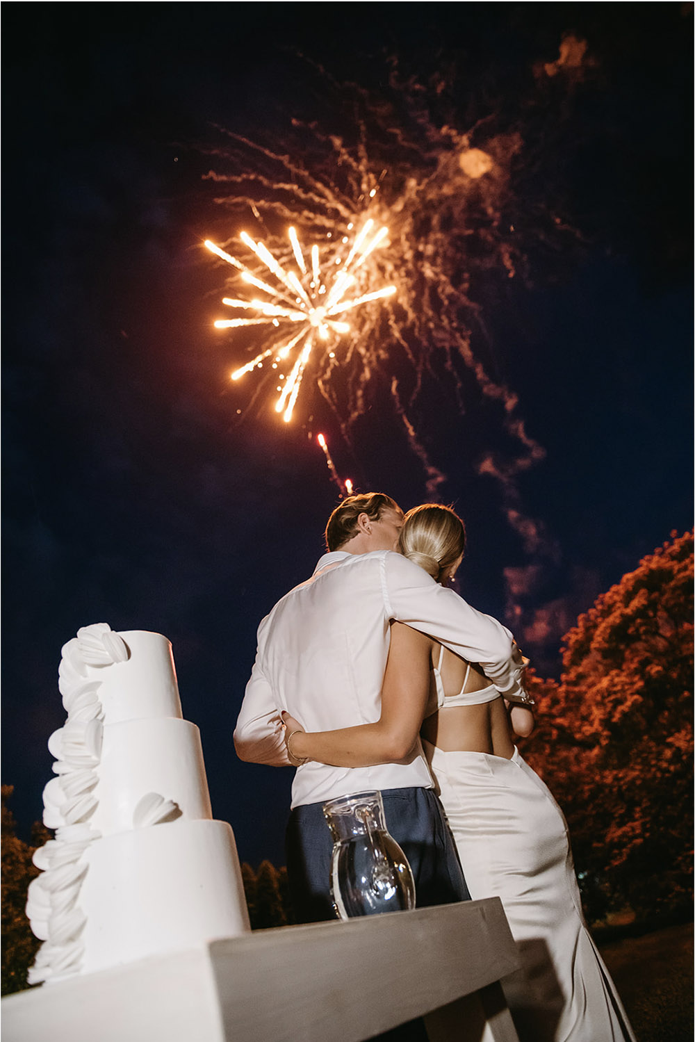 An editorial-style nighttime wedding portrait at an outdoor reception, showing a newlywed couple embracing beside a modern white wedding cake while fireworks burst overhead against the dark sky.