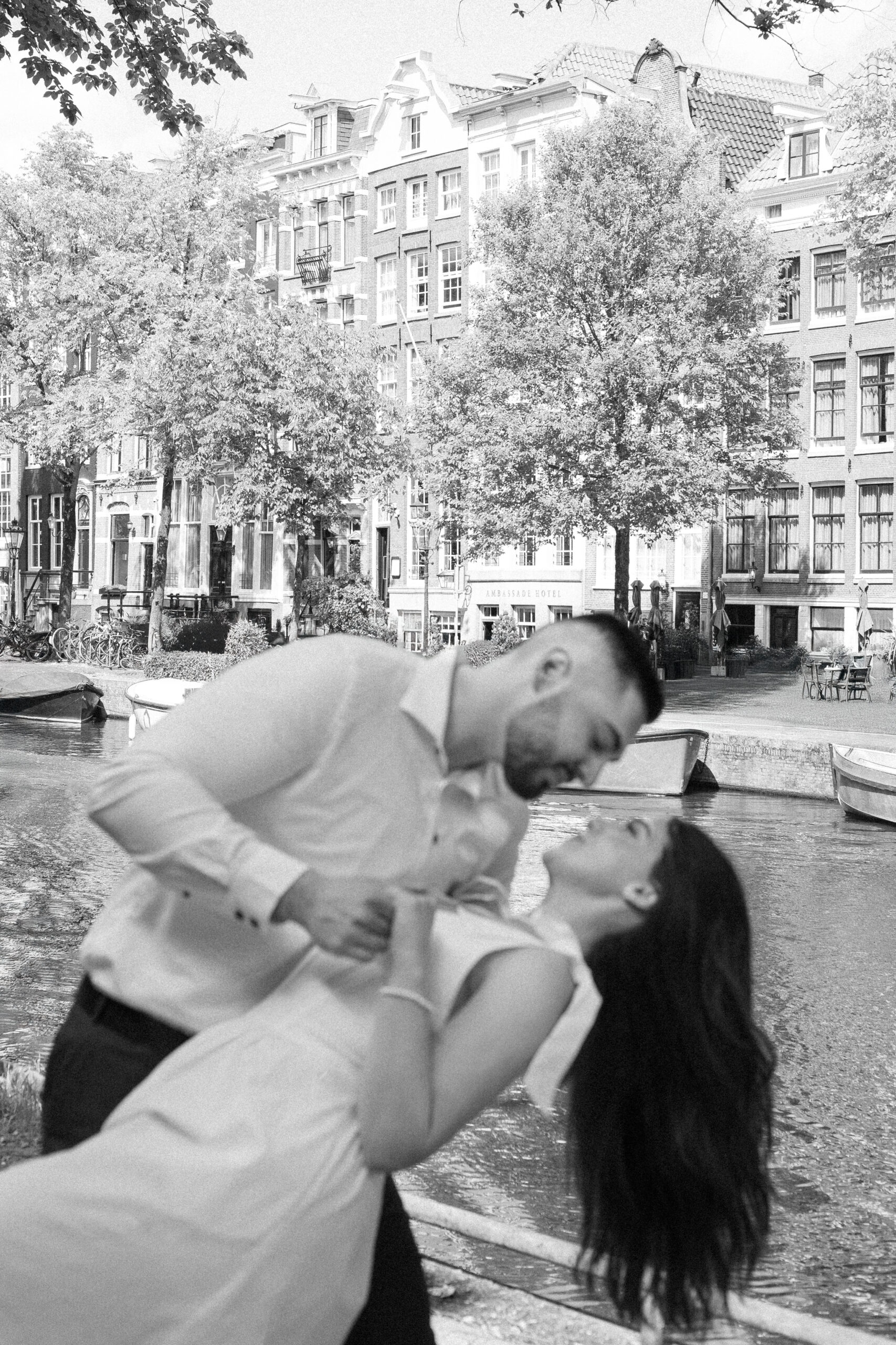 Black and white photo of a couple leaning for a kiss, with famous Amsterdam canal houses in the background