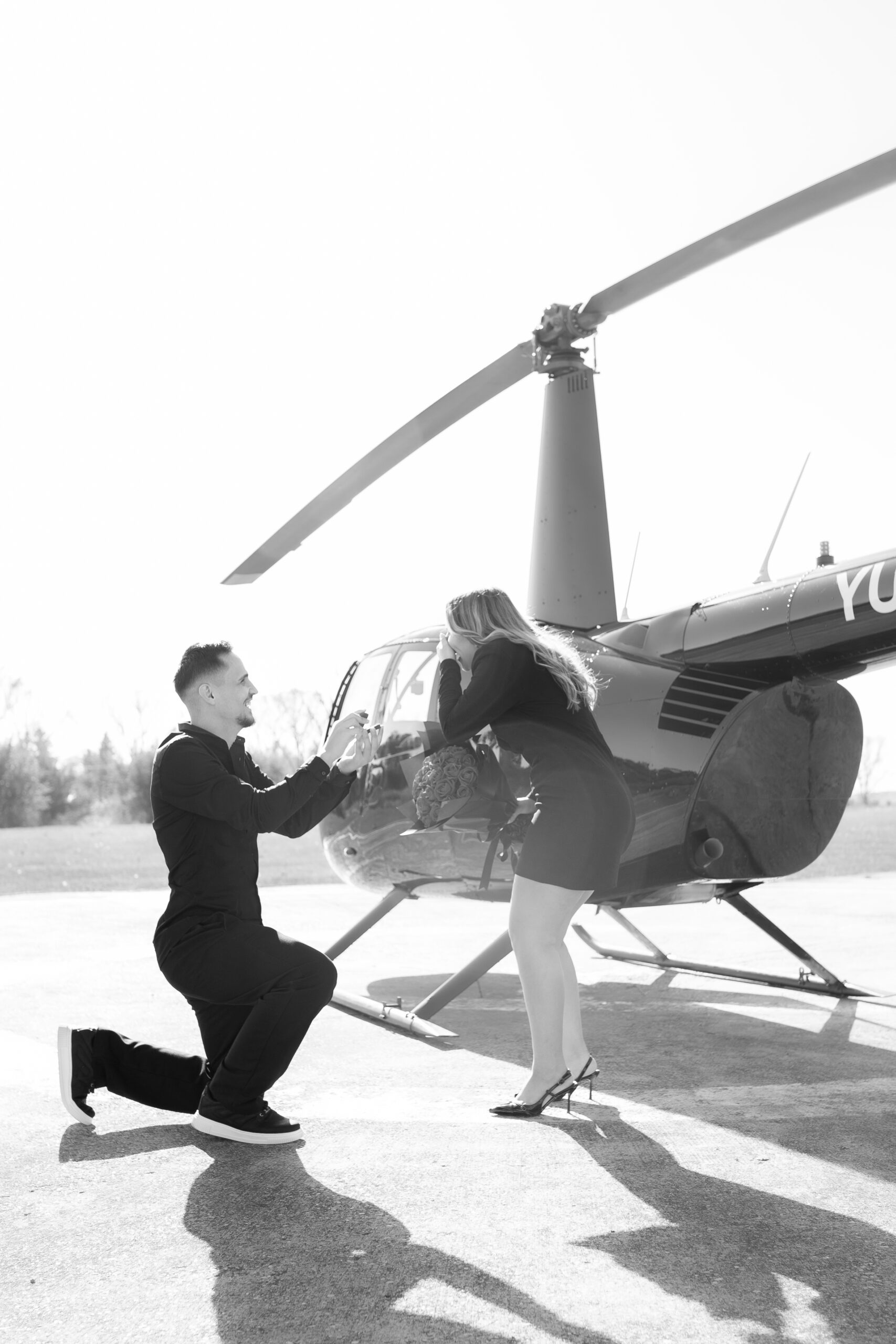 A person kneeling in front of their partner beside a helicopter, holding out a ring in a proposal moment.