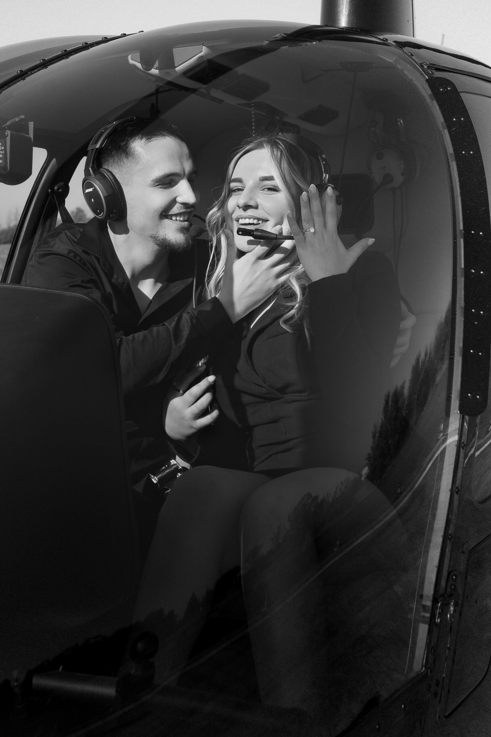 A black-and-white close-up of a couple laughing inside a helicopter, with the woman holding up her engagement ring.