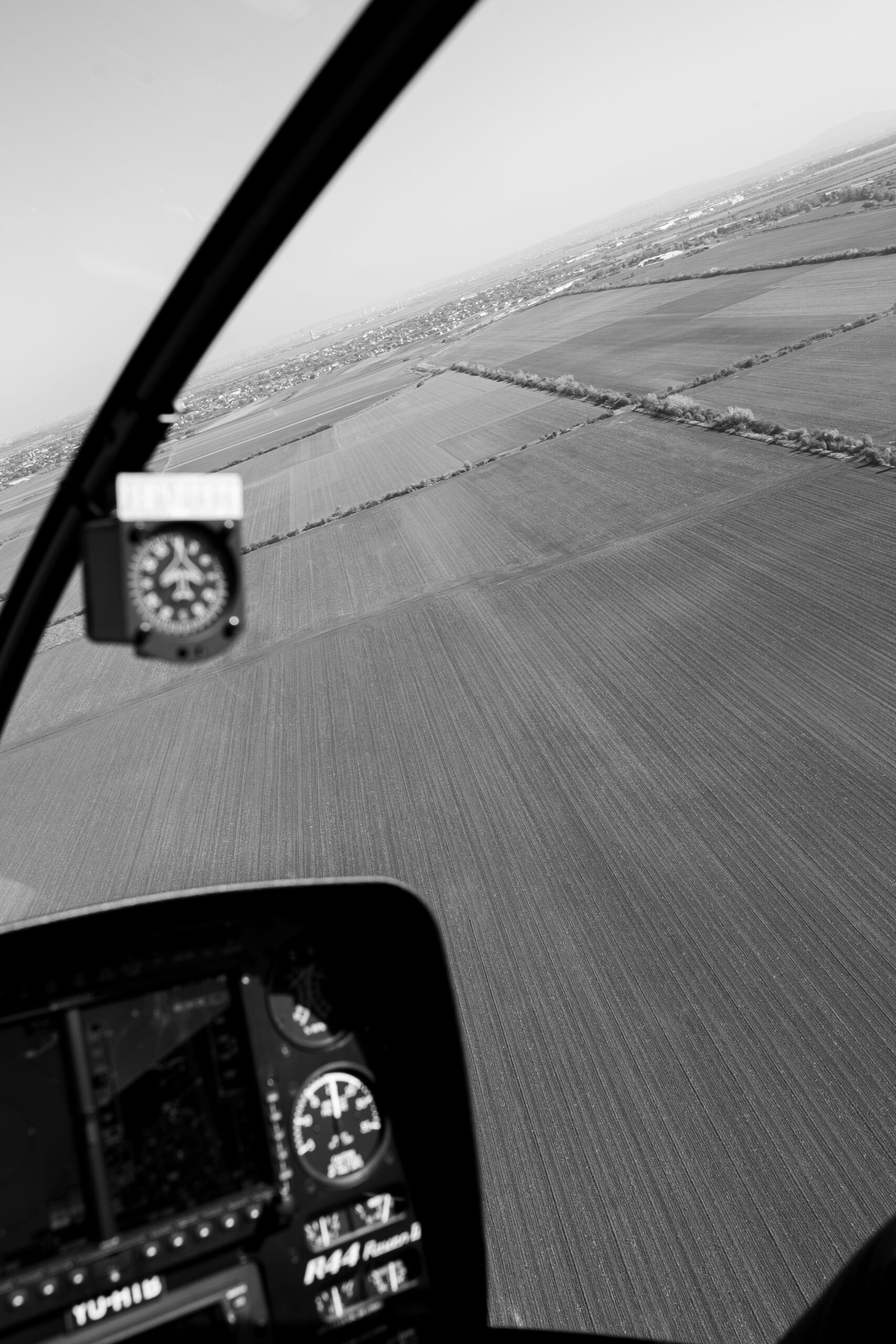 A black-and-white aerial view from inside a helicopter cockpit, looking out over open fields.