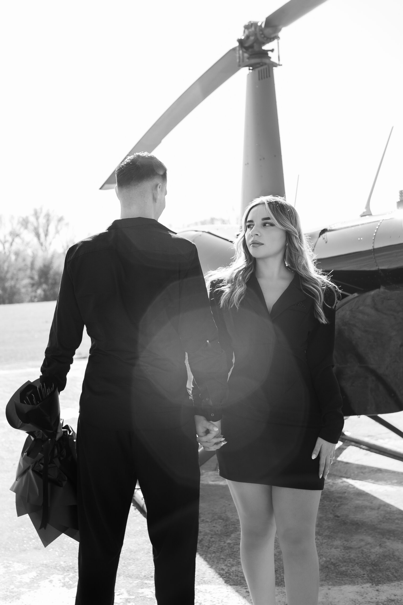 A black-and-white scene of a couple standing beside a helicopter, holding hands as the woman looks toward the camera.