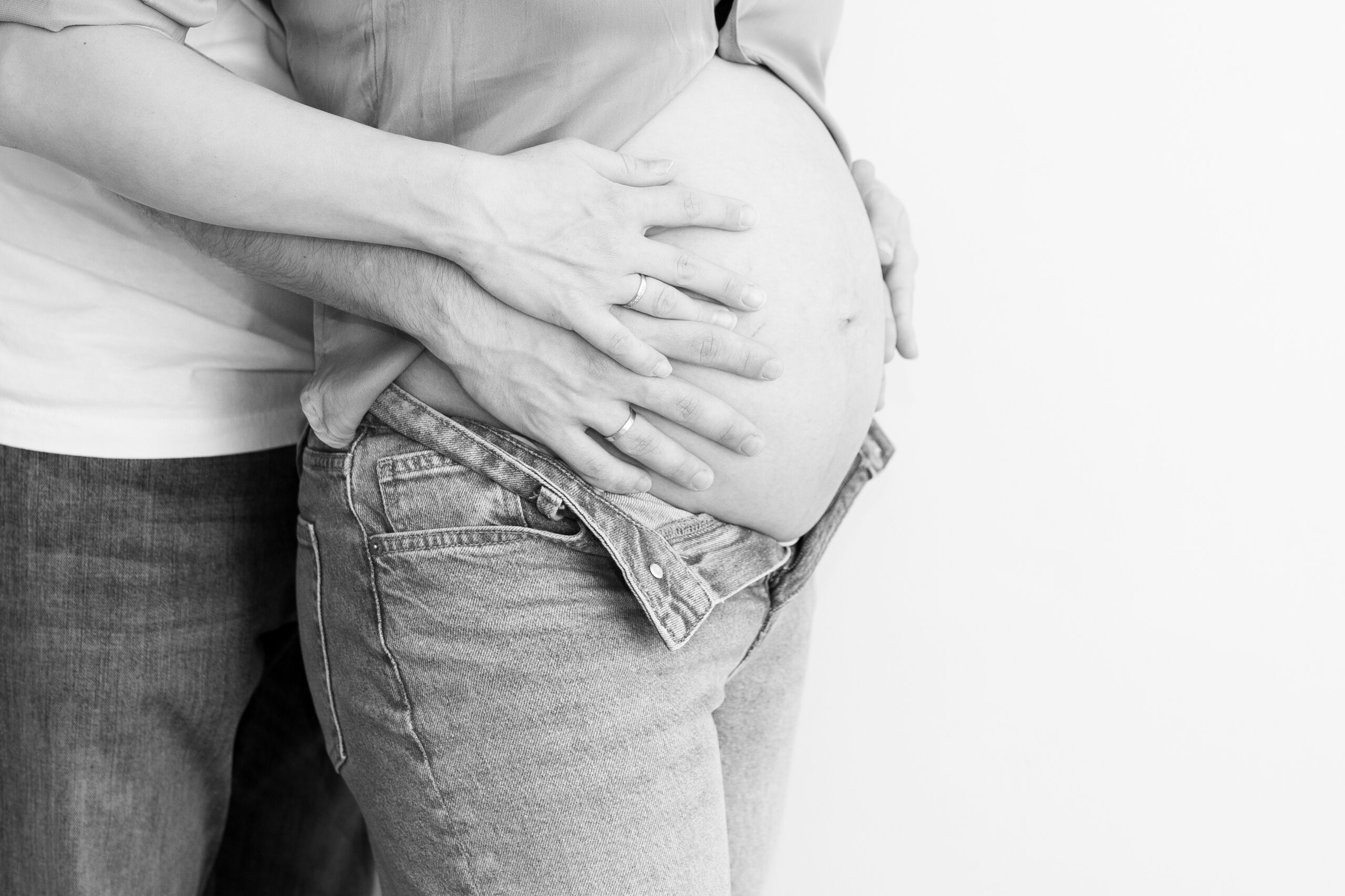 Trudnica i partner stoje zagrljeni, s rukama položenima na trudnički stomak, u crno-bijeloj fotografiji.