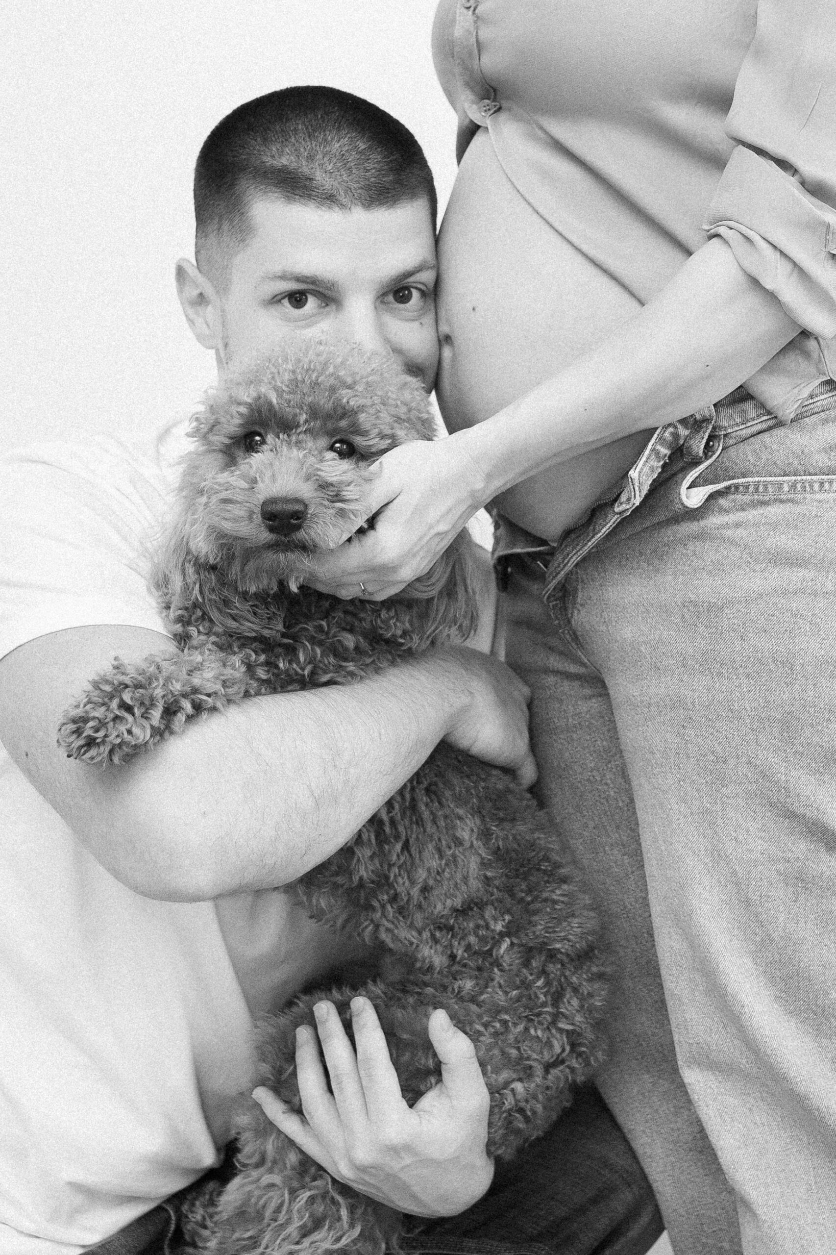 Par pozira sa psom, partner drži psa u naručju dok se oslanja uz trudnički stomak, crno-bijela fotografija.