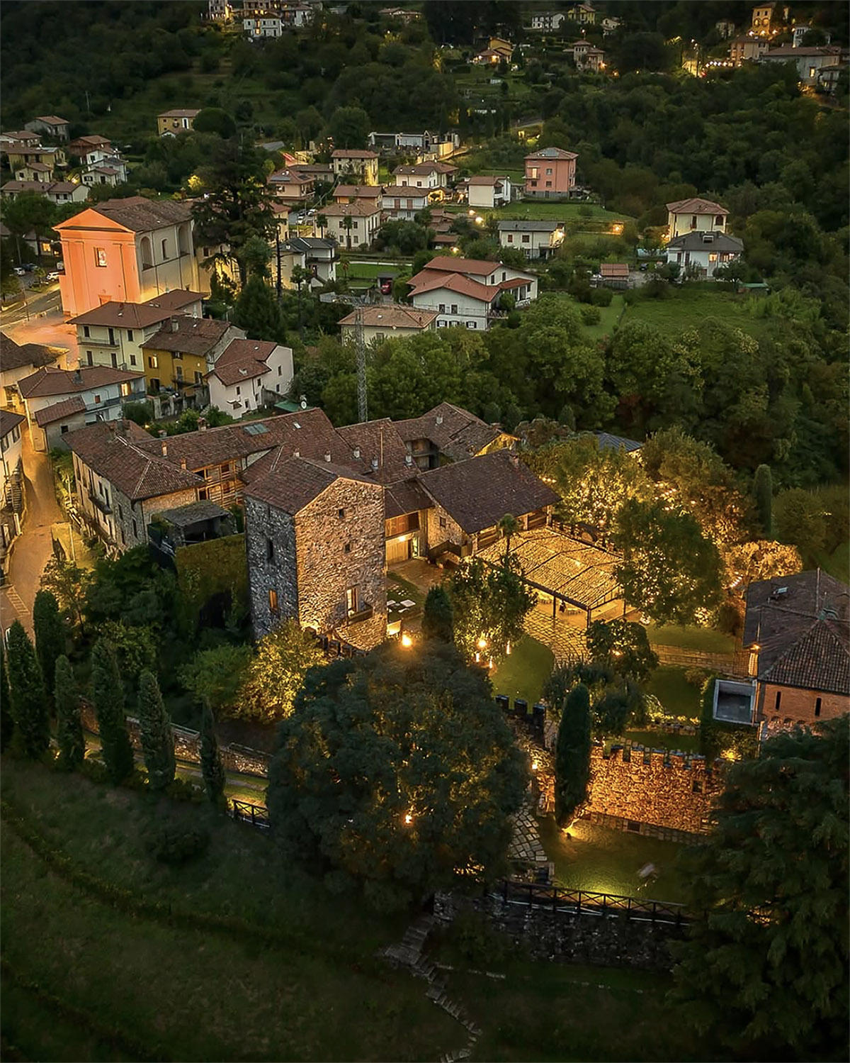 Castello di Rossino, Bergamo – aerial view of a hilltop wedding venue at dusk surrounded by glowing village lights and lush greenery in a cinematic editorial style.