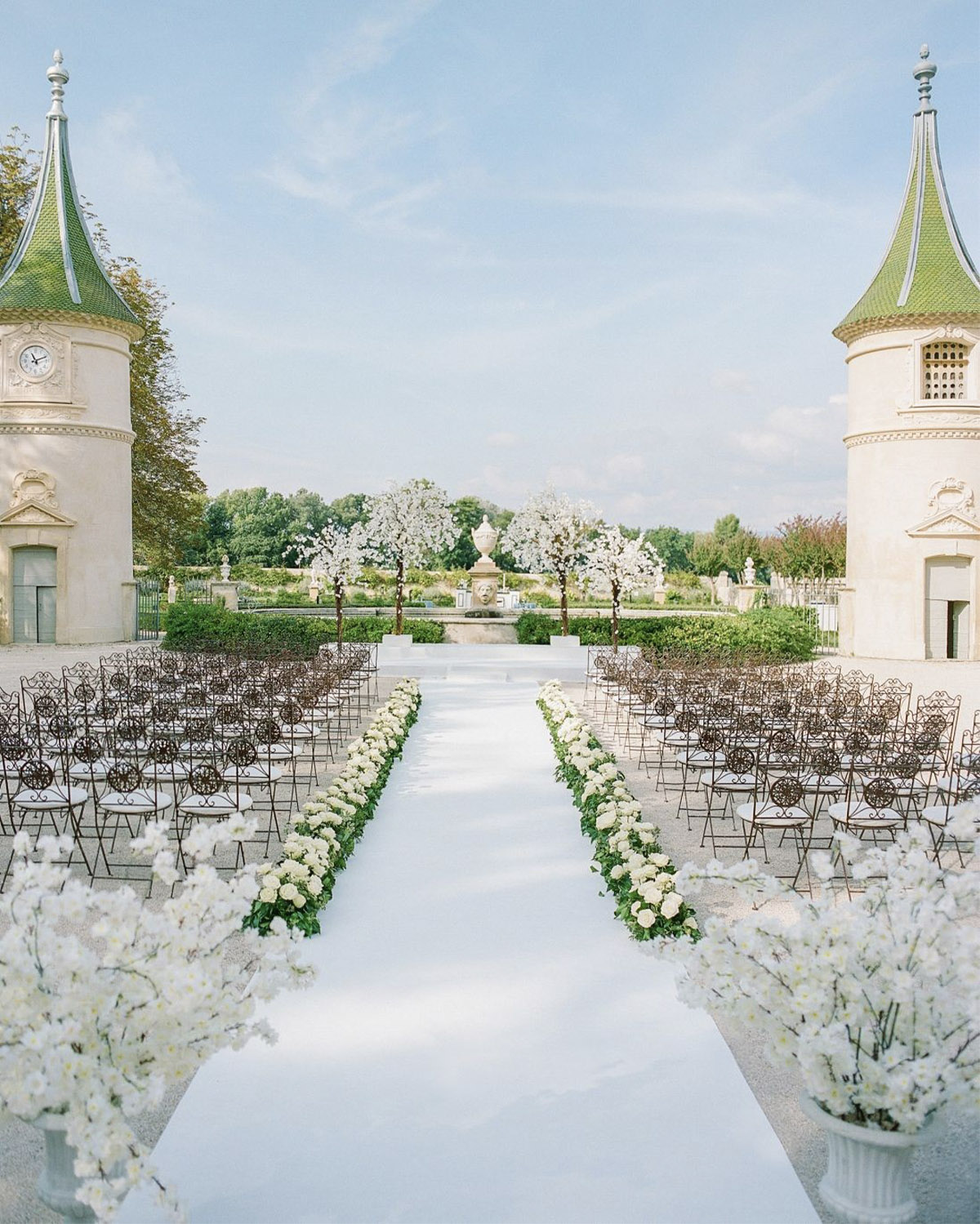 In Provence at a grand château, a symmetrical outdoor wedding aisle lined with white flowers leads between two ornate towers in a soft, light-filled editorial style.