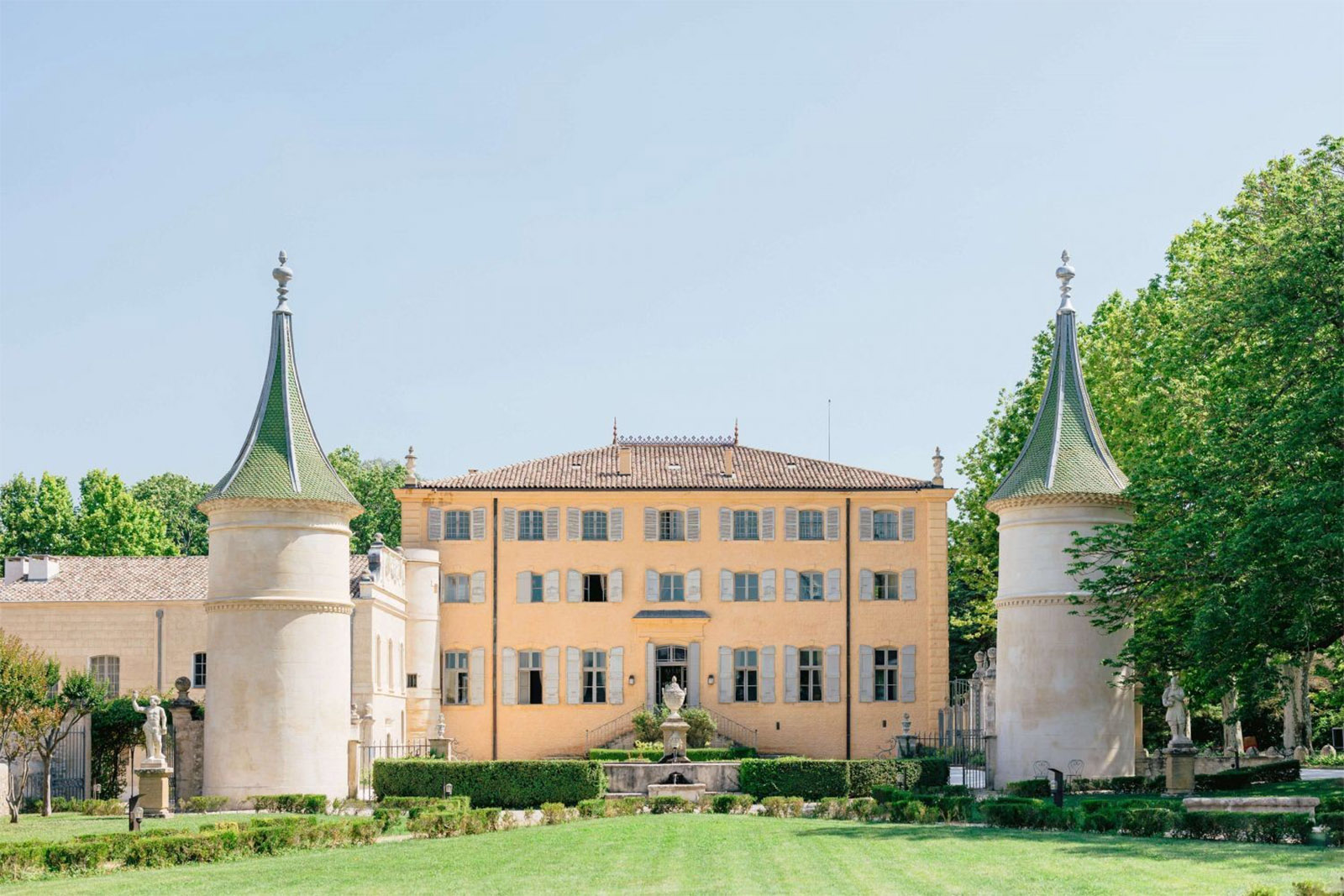 At a Provençal estate, a sunlit ochre château framed by two turreted towers sits beyond manicured gardens in an airy editorial style.