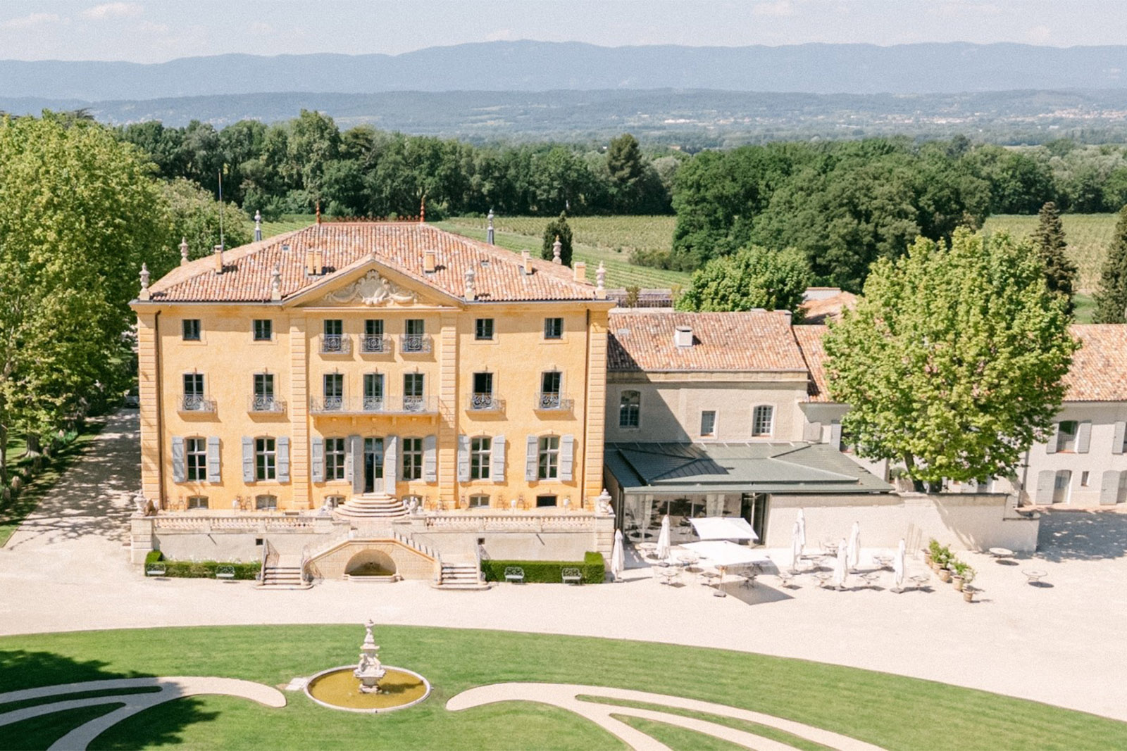 At a French estate, a sweeping view of the château façade and terrace overlooks manicured lawns in a sunlit editorial style.