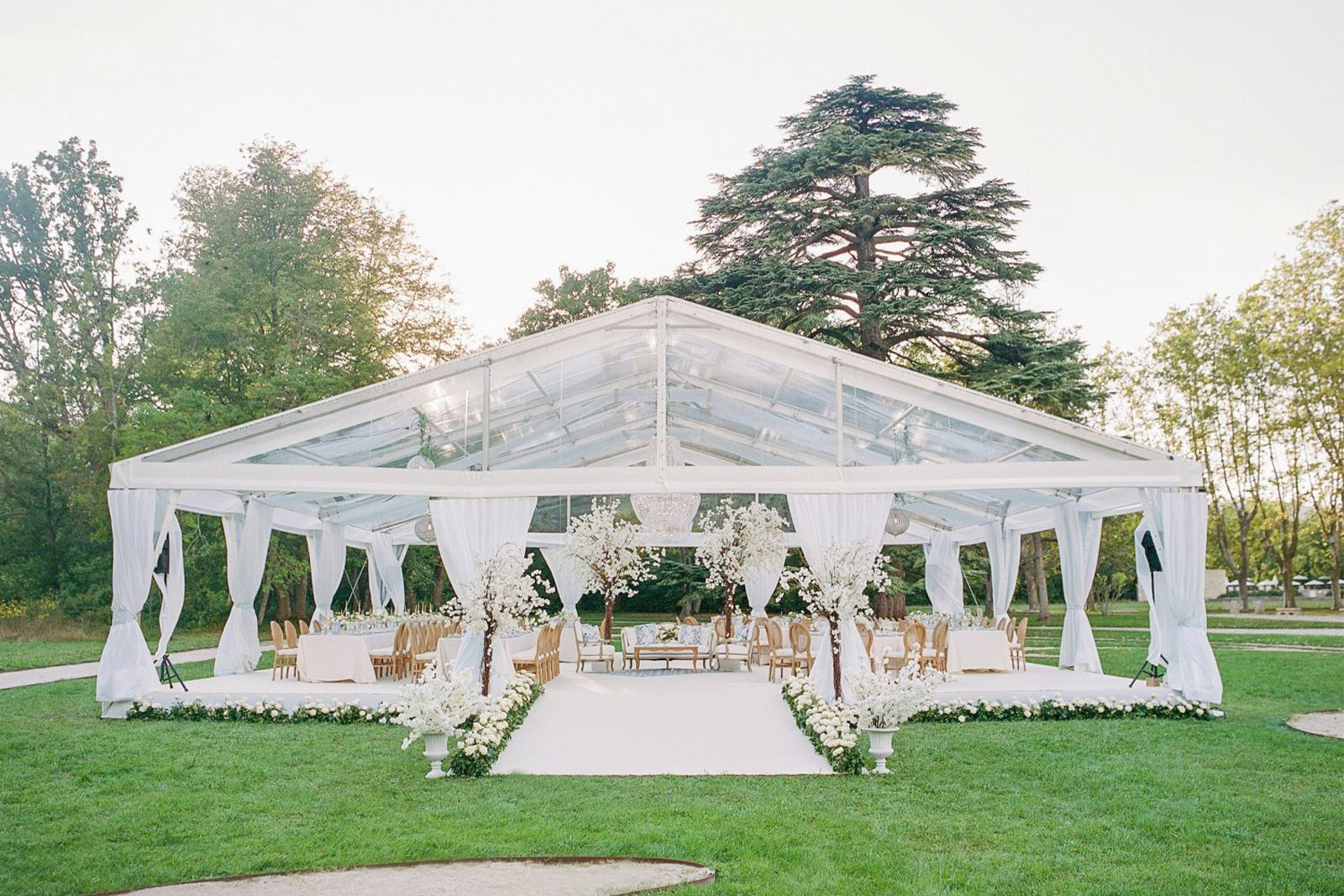 In Provence, a transparent reception tent with chandeliers and round tables is styled with white florals and greenery in a chic editorial wedding style.