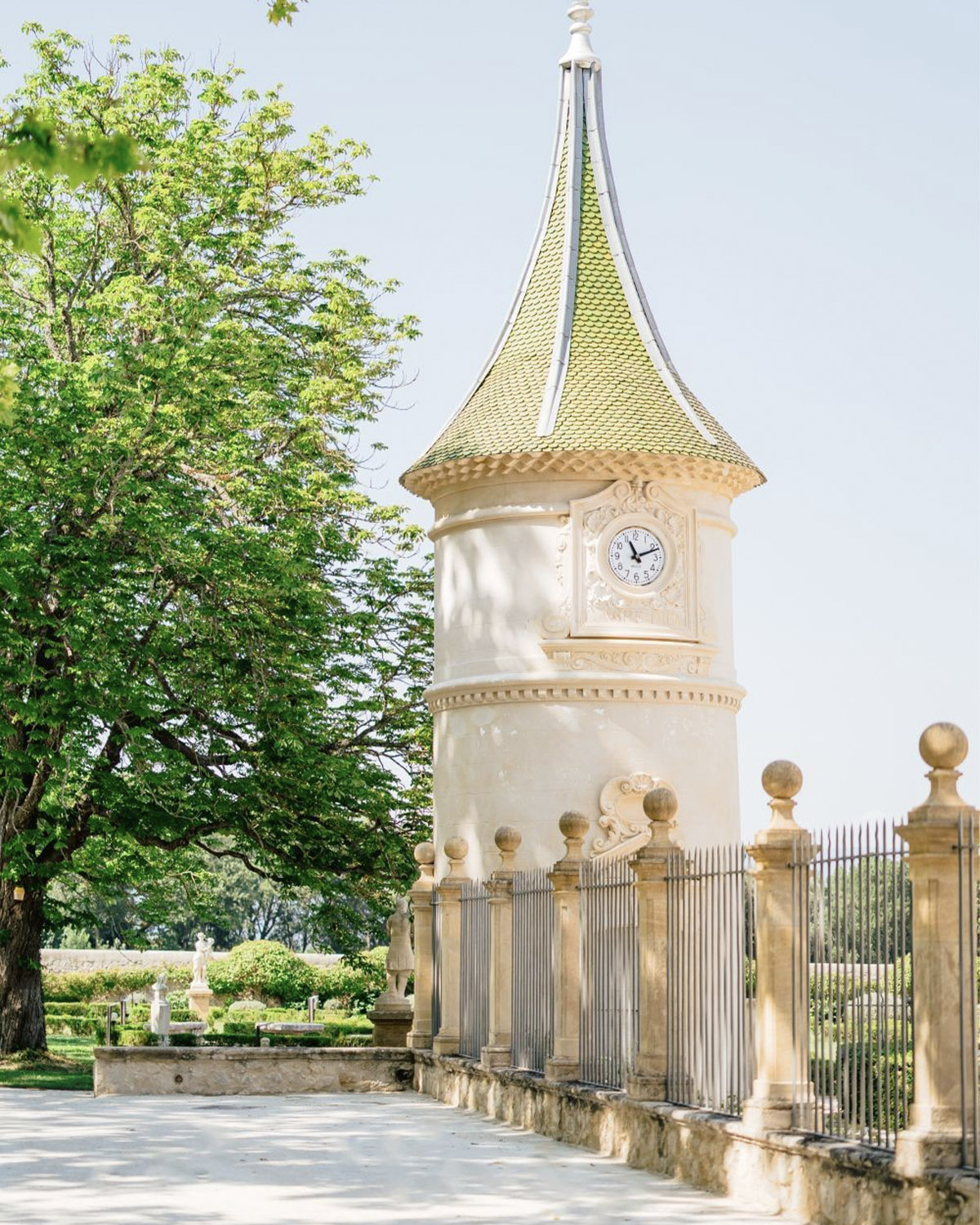In Provence, a close-up of a pale stone tower with a green-tiled spire and clock stands beside leafy trees in a bright, romantic editorial style.