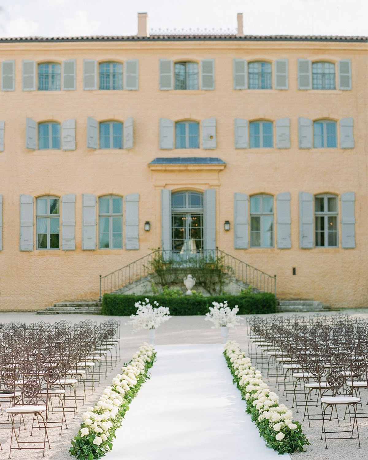 At a château in Provence, a floral-lined ceremony aisle with delicate white blooms faces a pastel façade in a refined editorial wedding style.