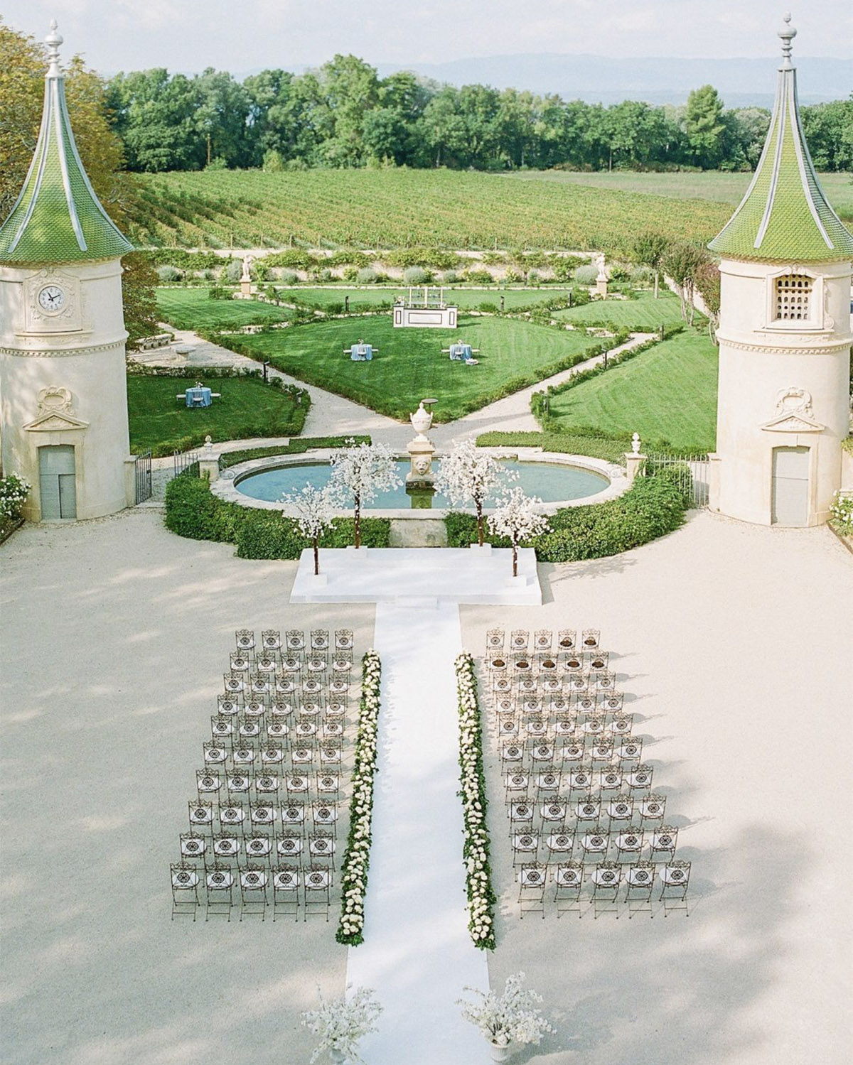In the gardens of a Provençal château, a bird’s-eye view shows a symmetrical ceremony setup with rows of chairs and a central aisle in a clean, editorial style.