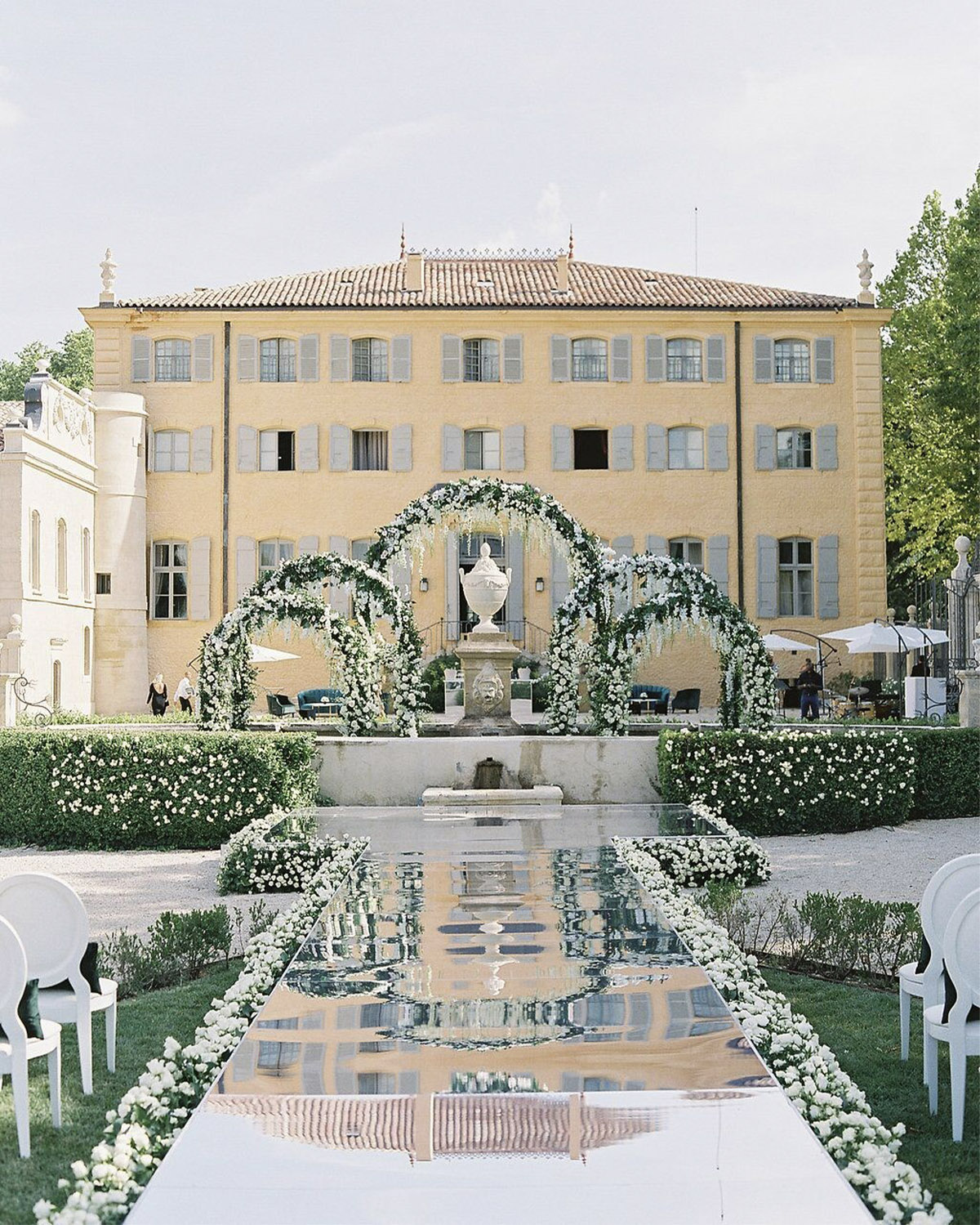 At a French countryside estate, a garden ceremony altar framed by fountains and greenery sits between two towers with vineyards beyond in an elegant editorial style.