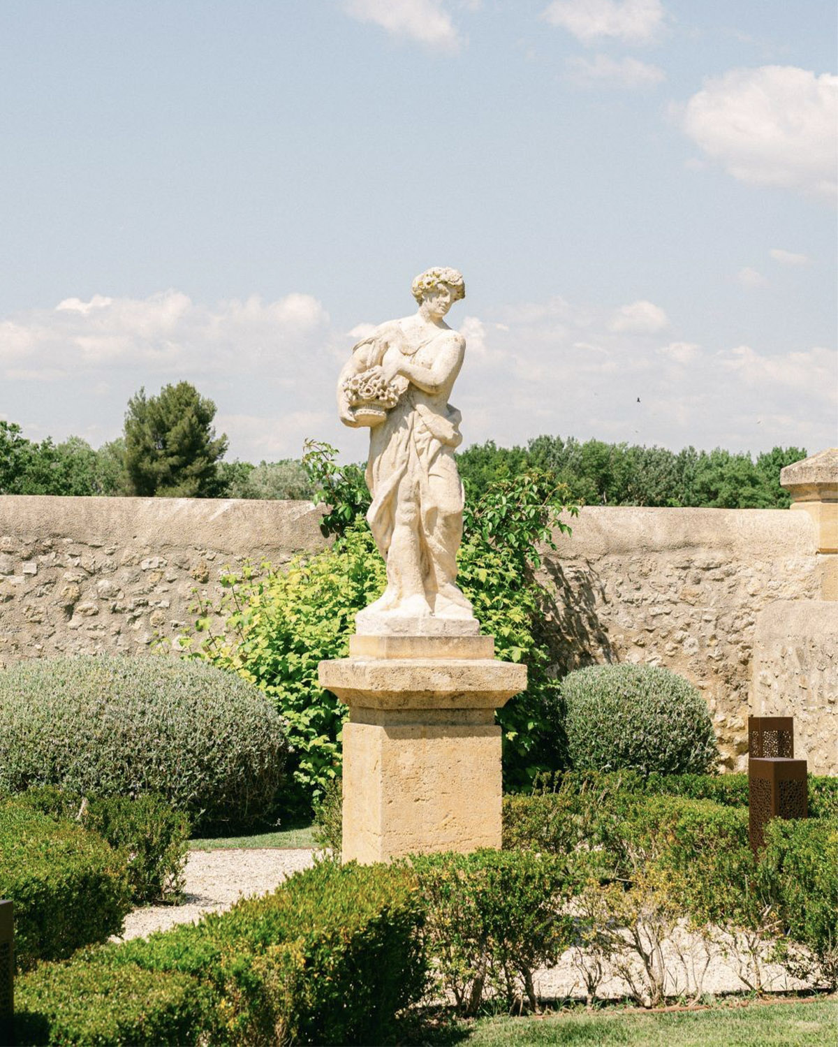 In a French château garden, a classical statue stands among trimmed hedges and stone walls under warm sunlight in a romantic editorial style.
