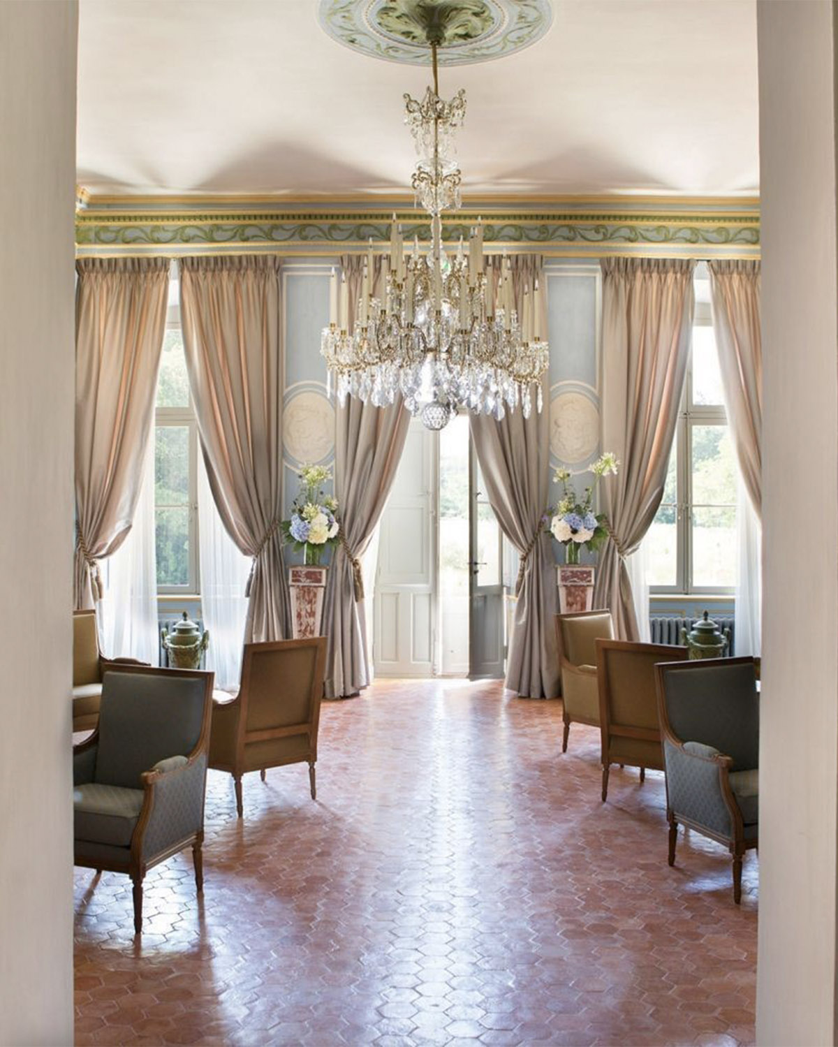 Inside a Provençal château, an elegant salon with crystal chandelier, tall draped windows, and antique seating is captured in a soft editorial style.
