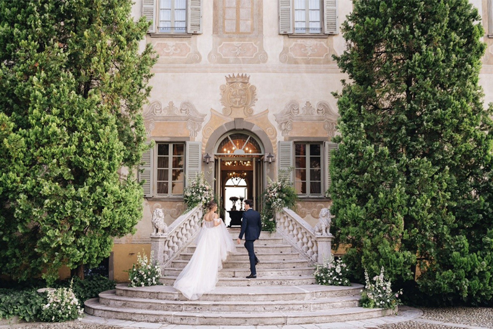 Bergamo, Italy historic villa entrance with a bride and groom walking up stone steps between manicured greenery, photographed in a romantic editorial wedding style.