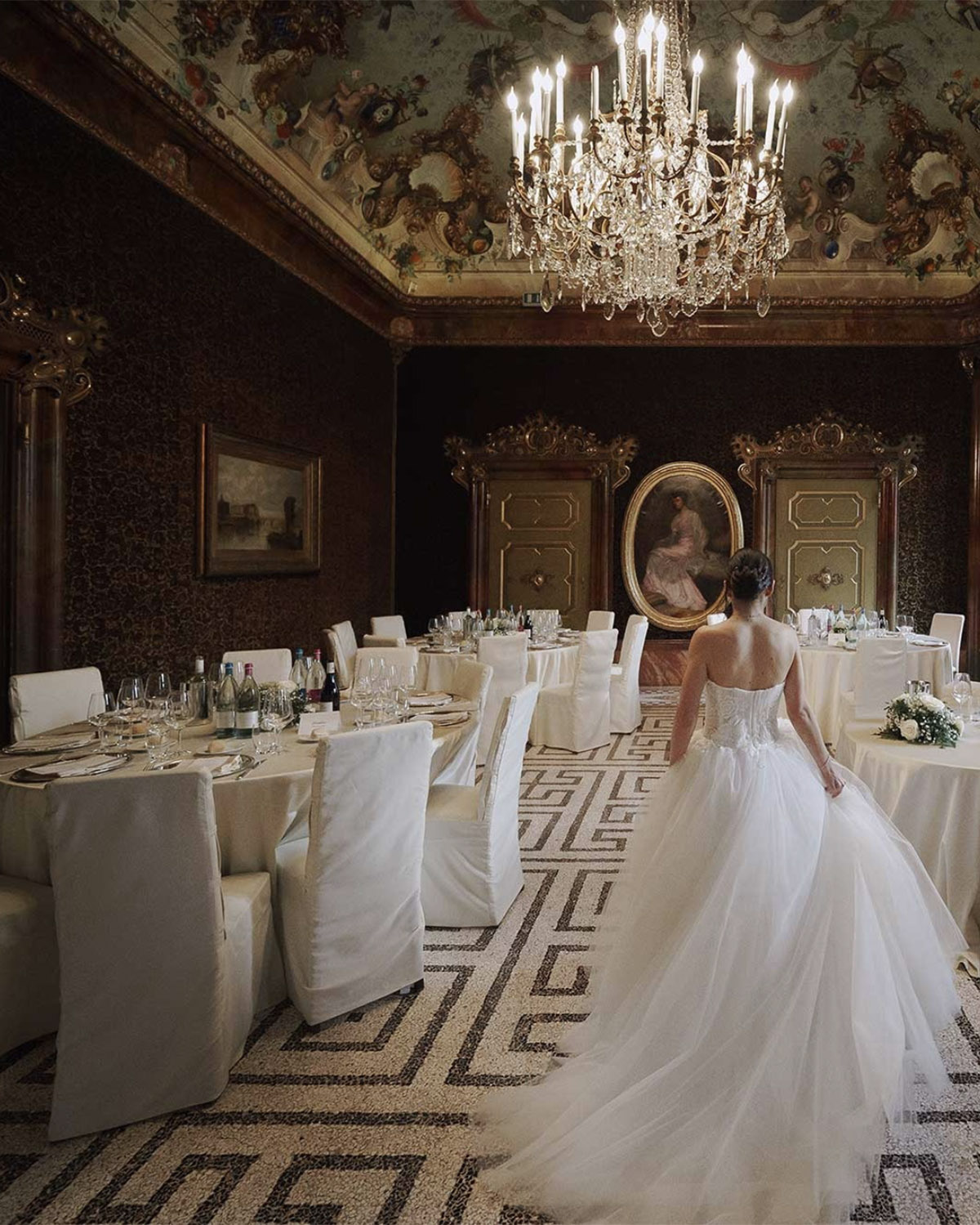 Bergamo, Italy opulent dining room with a bride walking among round tables under a chandelier, styled in a dramatic editorial wedding fashion aesthetic.