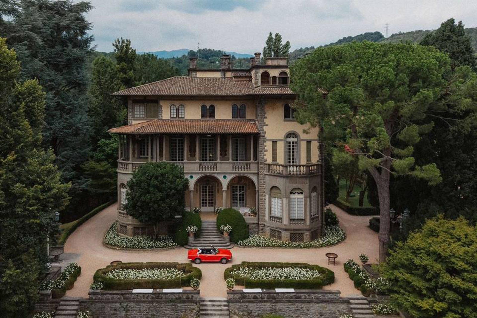 A historic Italian villa in Bergamo sits surrounded by lush greenery and rolling hills, with a small red convertible parked out front, captured in a refined editorial style.