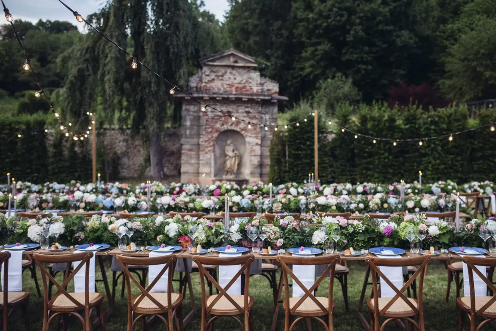 In a garden courtyard in Bergamo, long banquet tables are decorated with abundant pastel florals and glassware beneath string lights, styled in a romantic editorial aesthetic.