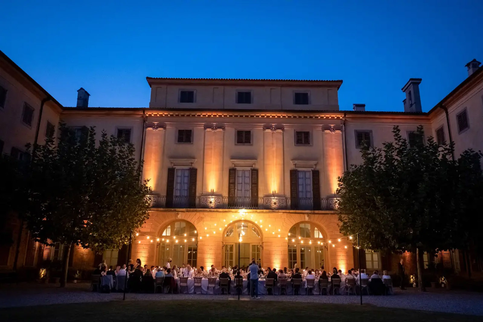A grand villa in Bergamo glows at dusk as guests dine at a long table in the courtyard, captured in a cinematic editorial style.