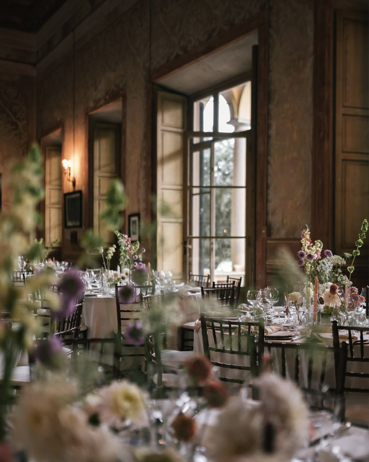 Inside an elegant villa in Bergamo, round reception tables are set with delicate florals and glassware near tall windows, photographed in a warm, fine-art editorial style.