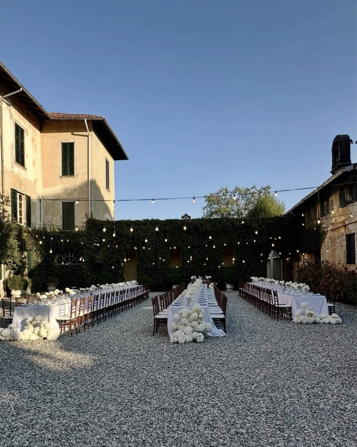 A sunlit courtyard in Bergamo features long reception tables with white florals and string lights overhead, styled in a clean editorial aesthetic.