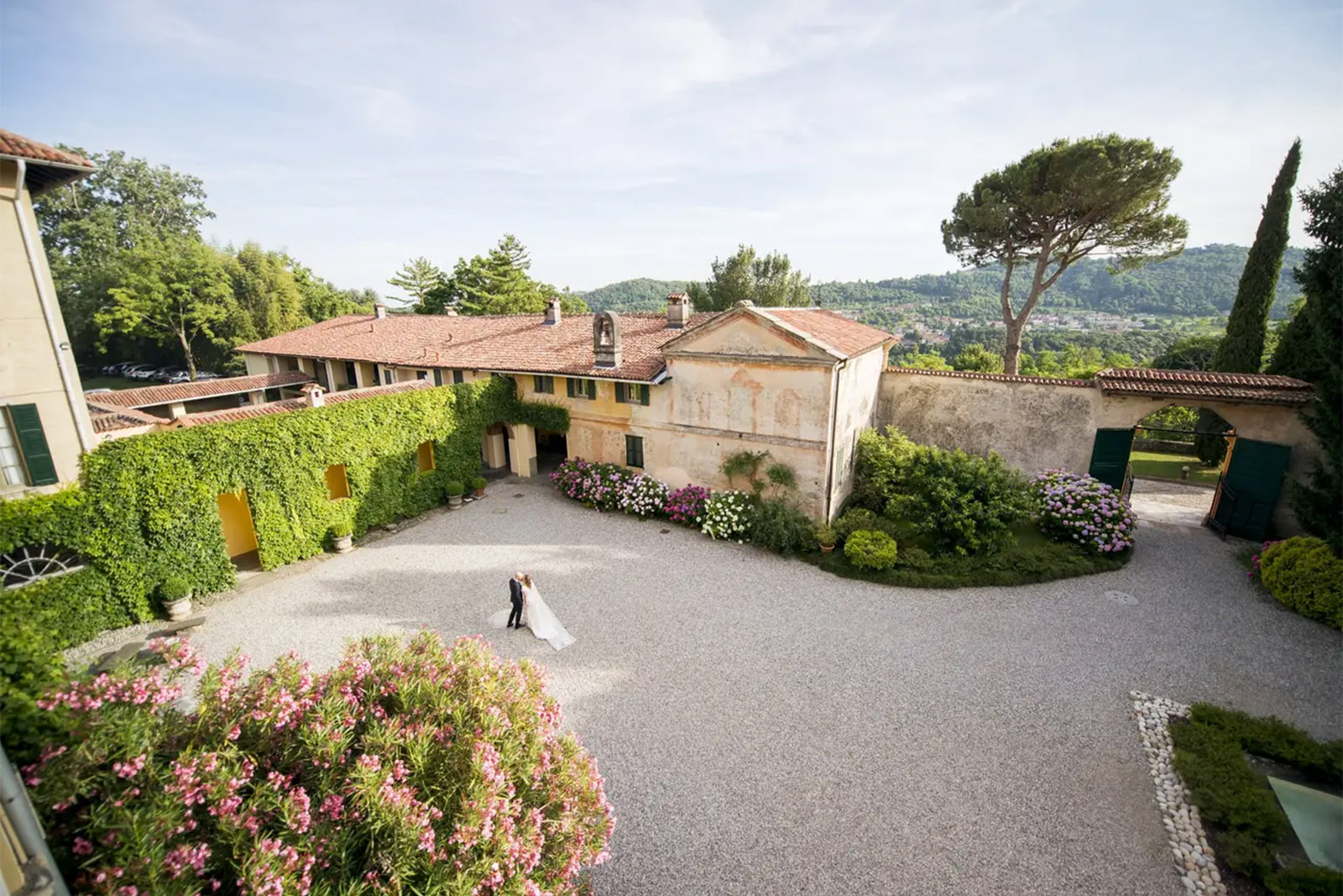 In a countryside villa courtyard in Bergamo, a couple walks across a gravel space framed by ivy-covered walls and blooming flowers, captured in a romantic editorial style.
