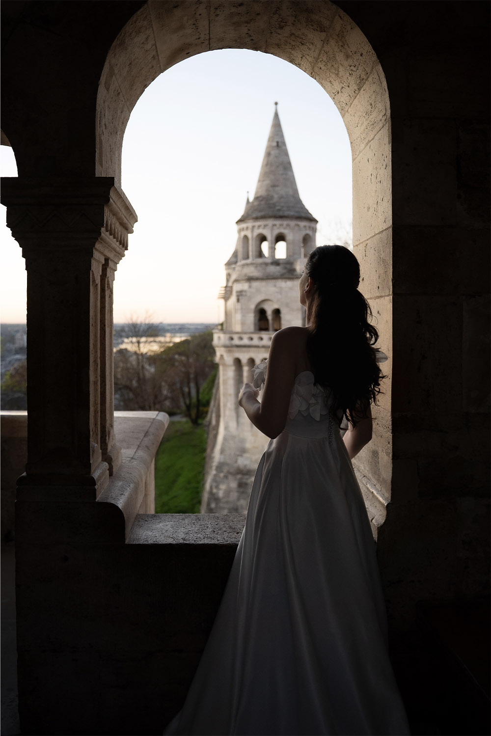 At Fisherman’s Bastion in Budapest, a bride in a flowing white gown stands framed by a stone archway, gazing toward a fairytale turret in a soft, romantic editorial style.