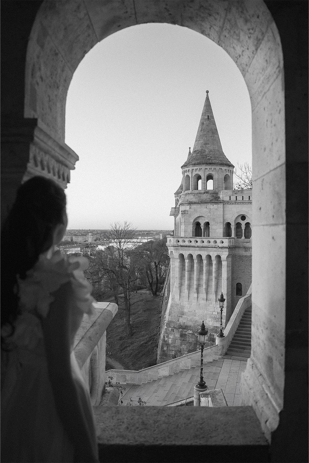 At Fisherman’s Bastion in Budapest, a softly blurred bride looks out from an arched balcony toward a historic tower, captured in a timeless black-and-white editorial style.