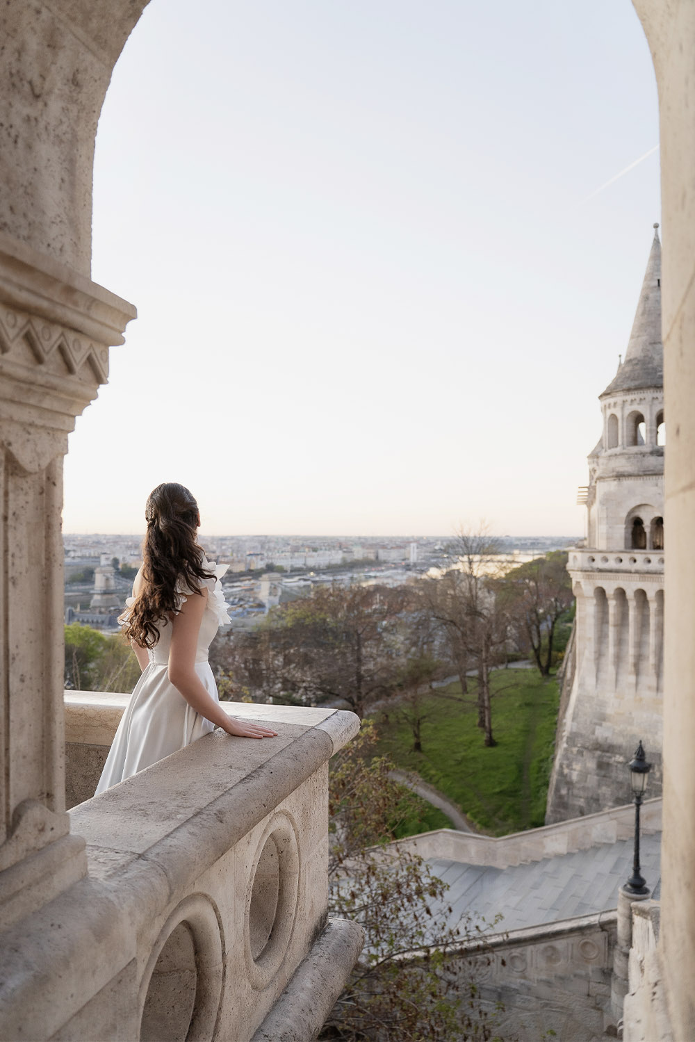 At Fisherman’s Bastion in Budapest, a bride in a white dress leans on a balcony overlooking the city and river at sunset, portrayed in a light, airy editorial style.