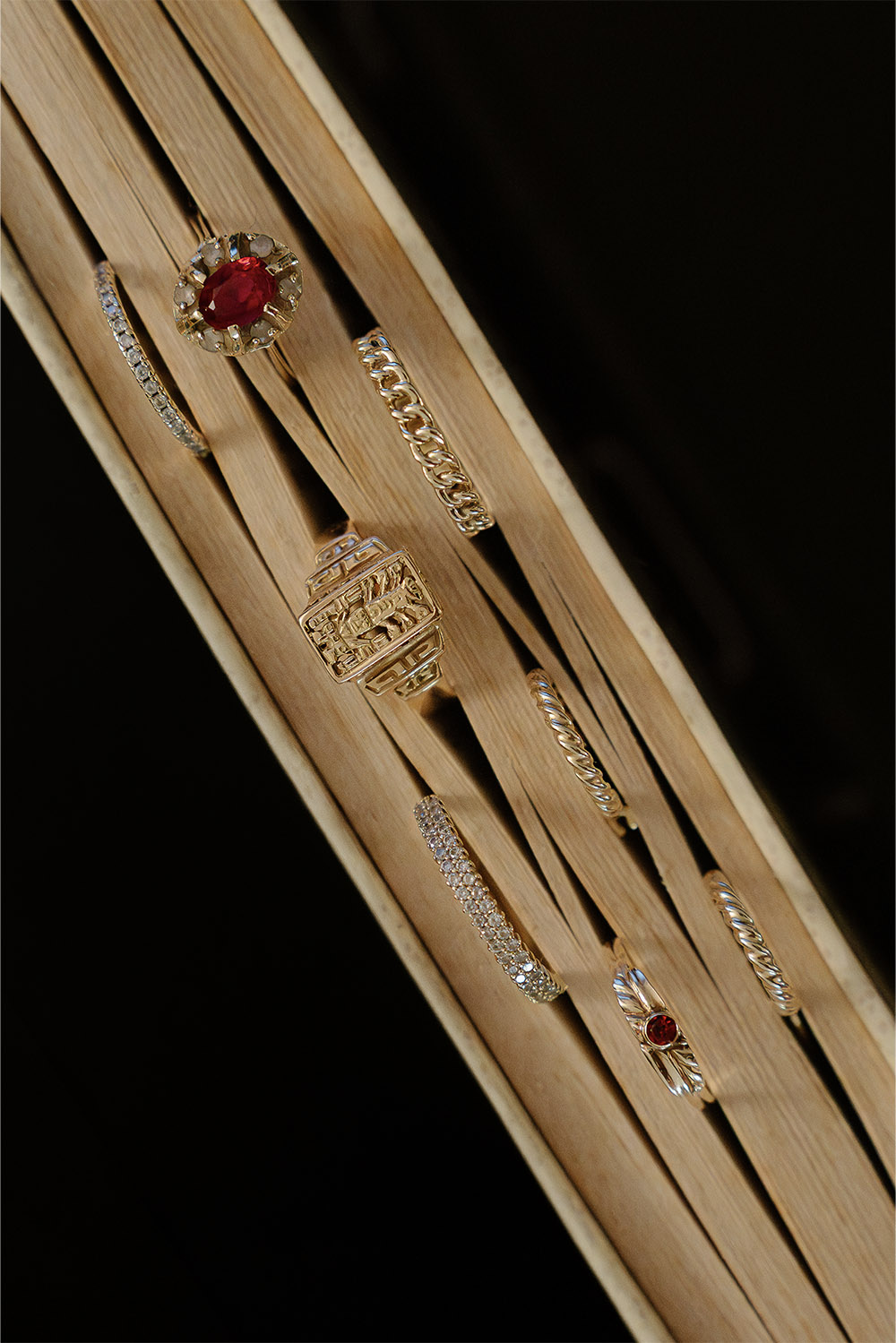 In a studio setting, a collection of gold rings with diamonds and red gemstones rests on wooden surfaces, photographed in a rich, high-contrast editorial jewelry style.