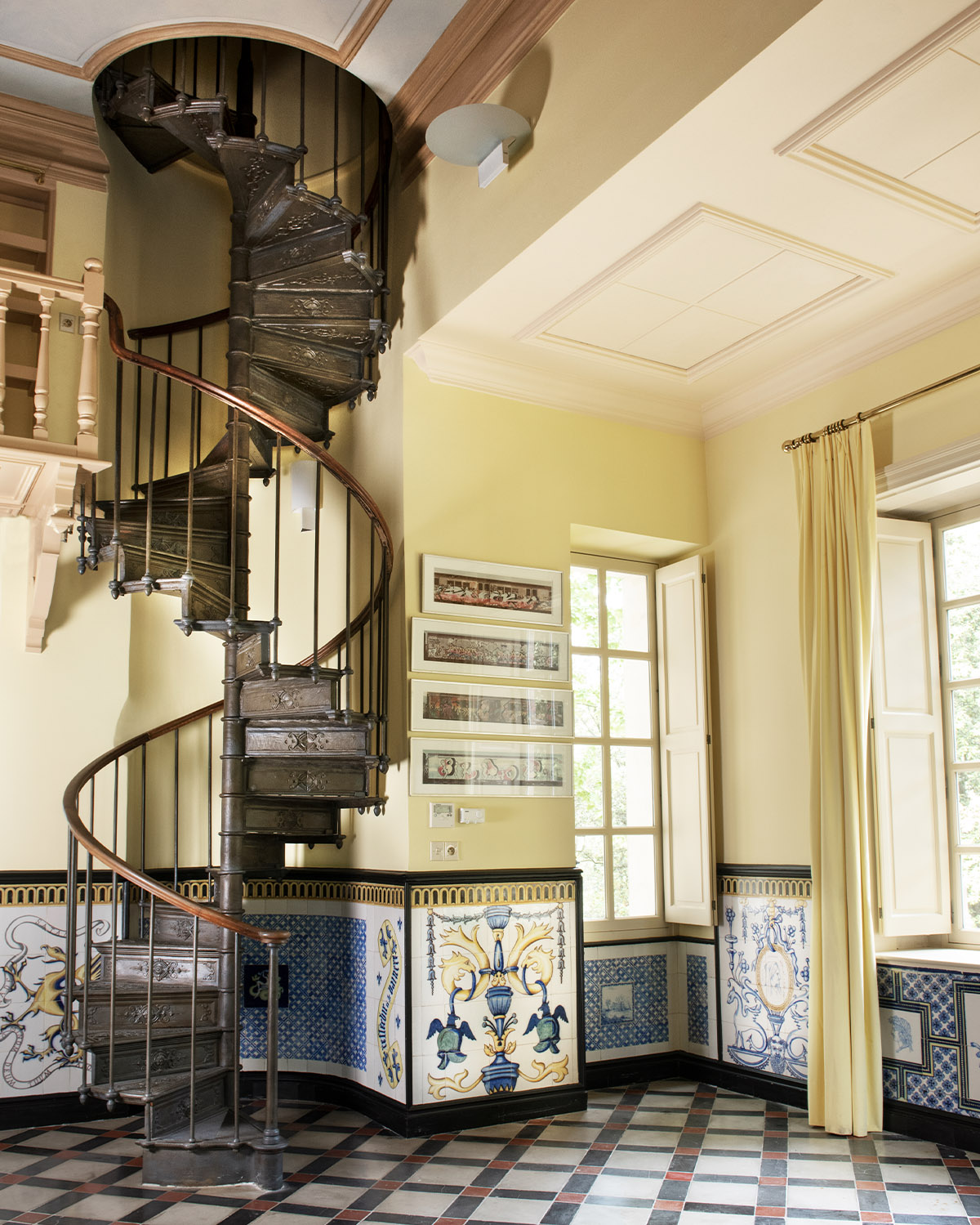 Inside a Provençal château, a dark wrought-iron spiral staircase curves upward beside sunlit windows and decorative ceramic wall panels, photographed in a warm, elegant interior editorial style.