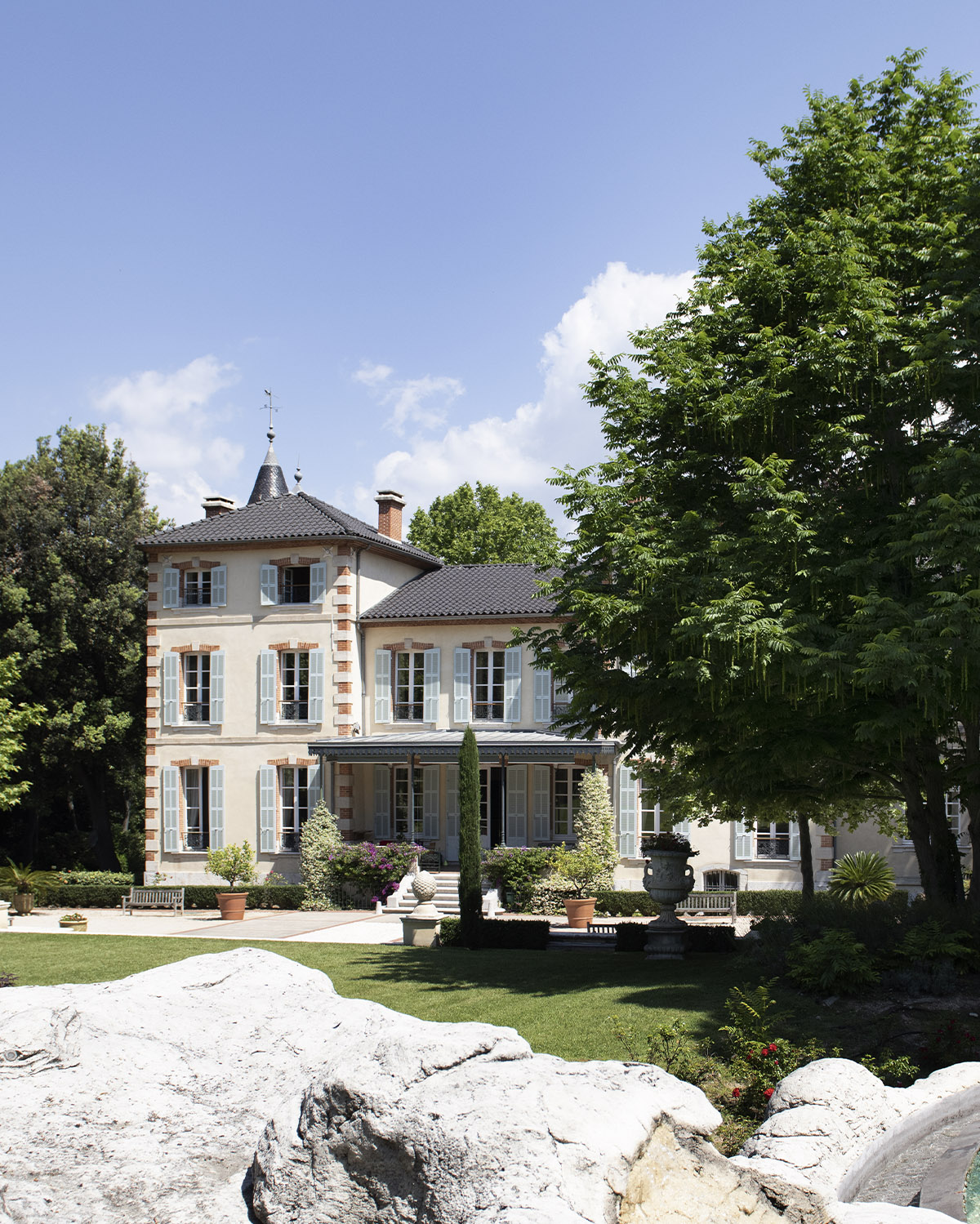 A sunlit view of Château de Forbin in Provence, France, showing a pale stone manor framed by lush trees and manicured gardens in a refined editorial style.