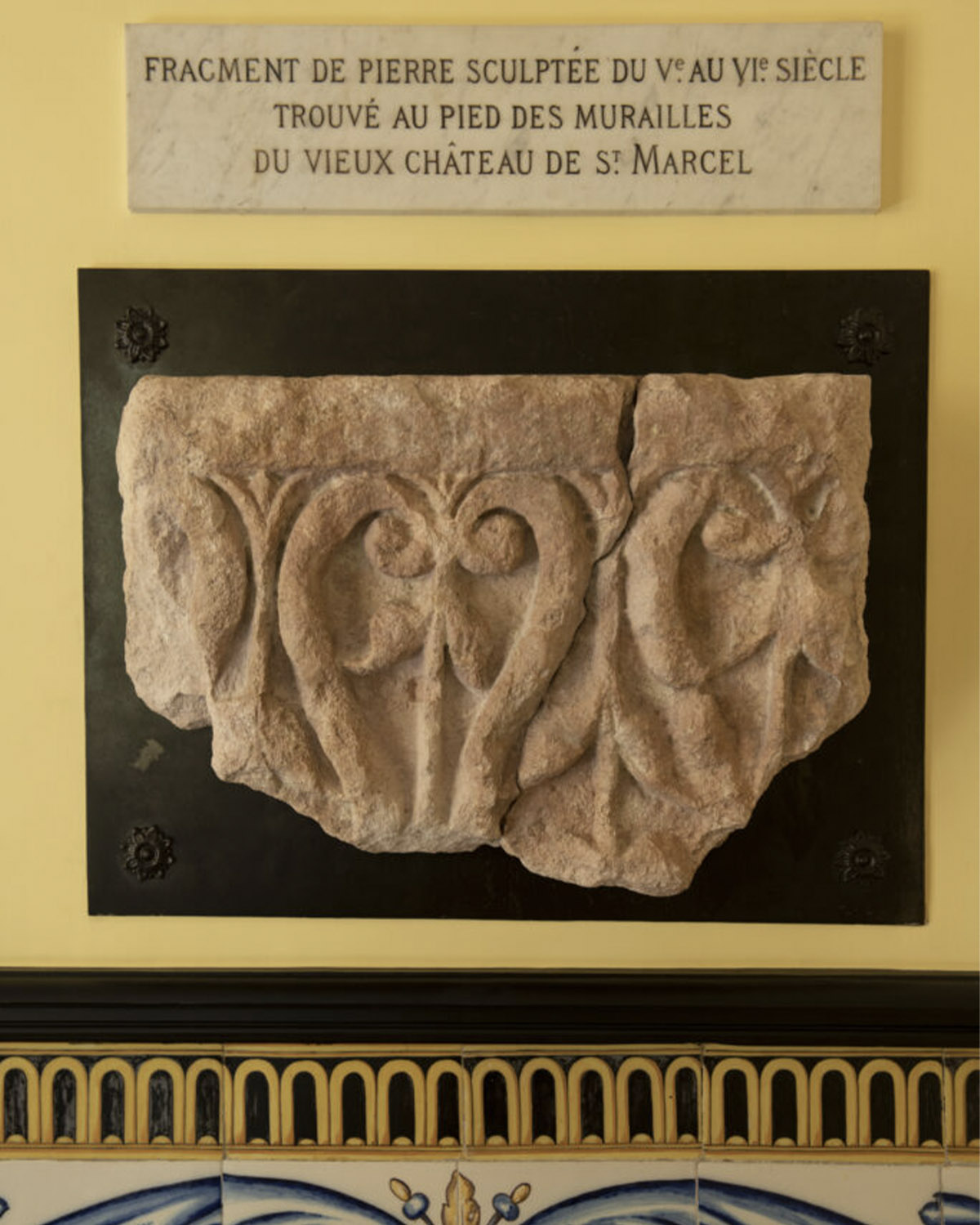 In a château interior in southern France, a weathered carved stone fragment is mounted beneath a French inscription plaque, presented in a museum-like editorial style emphasizing historical texture and detail.
