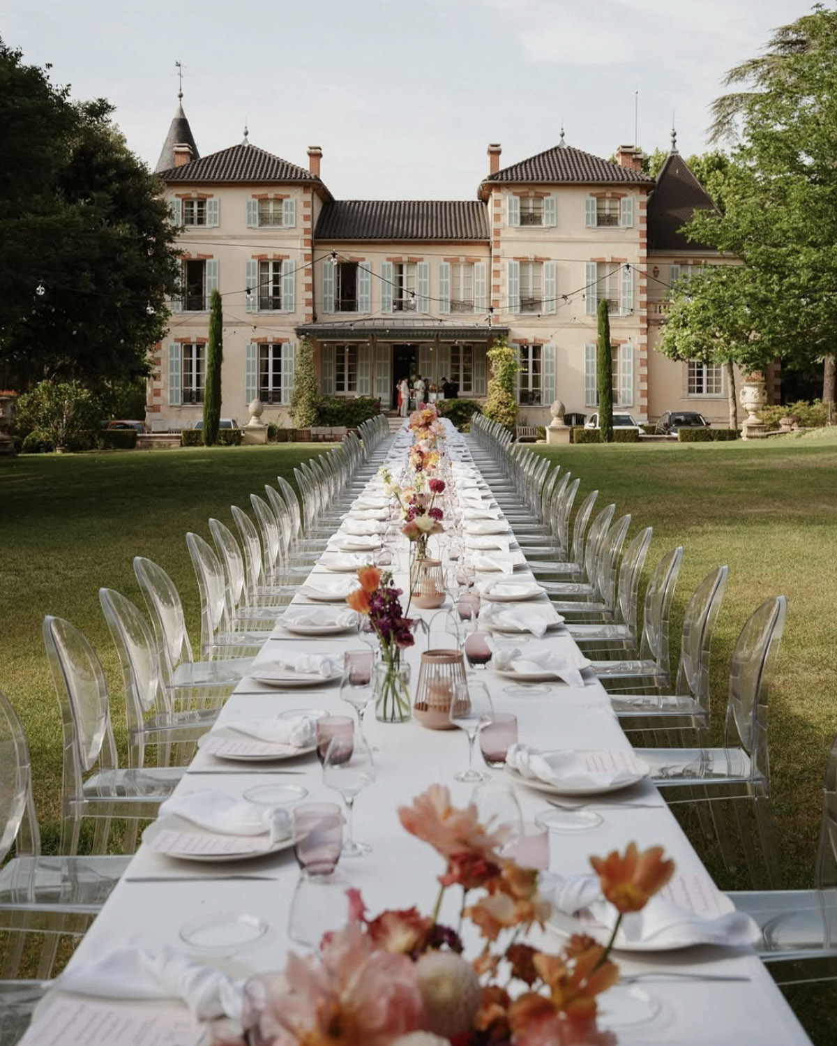 At Château de Forbin in Provence, France, a long outdoor wedding table lined with transparent chairs and soft floral arrangements stretches across the lawn toward the elegant estate in a romantic editorial style.