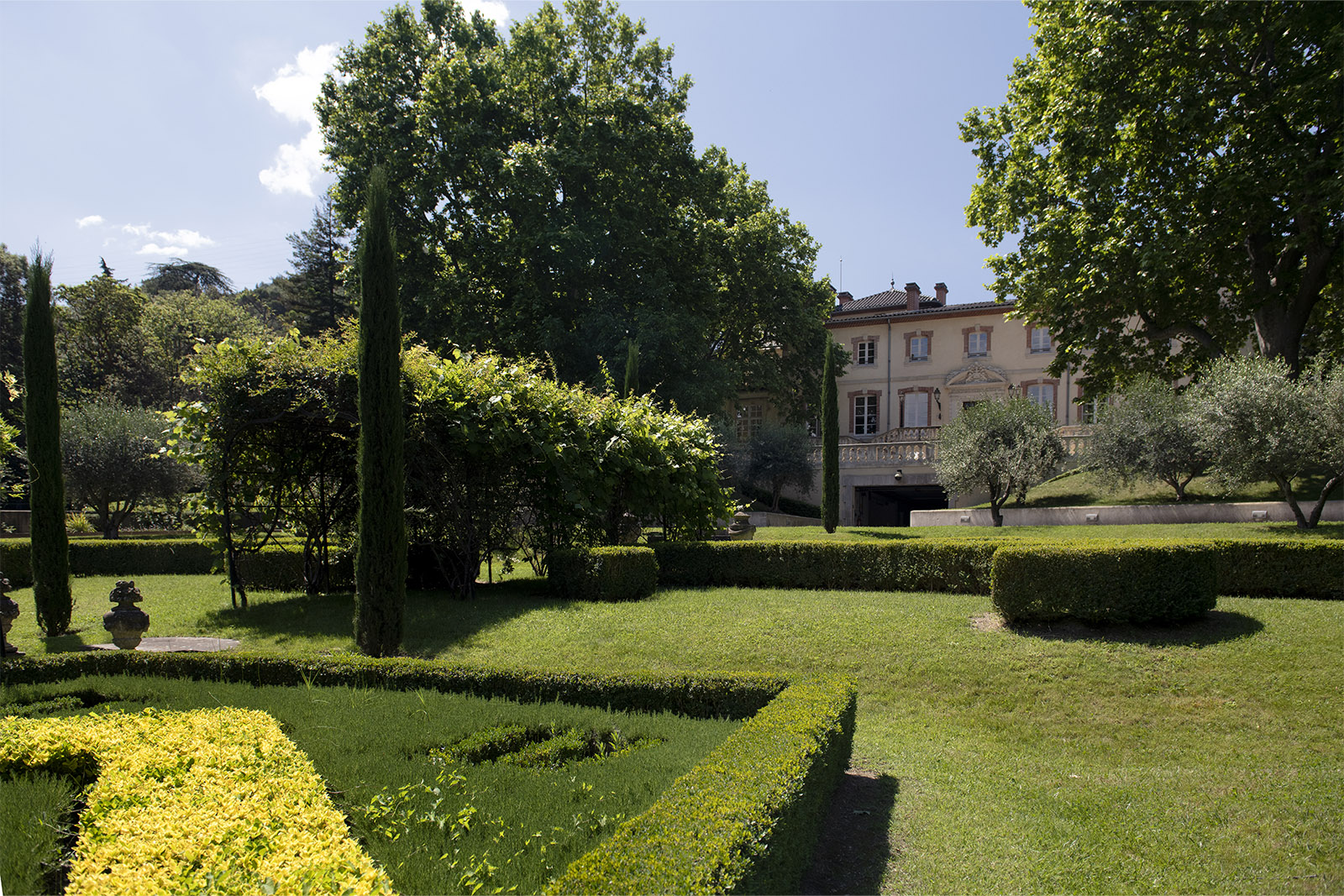 In the landscaped gardens of a Provençal estate, manicured hedges and tall trees frame a distant château under bright sunlight, shown in a relaxed, scenic editorial style.