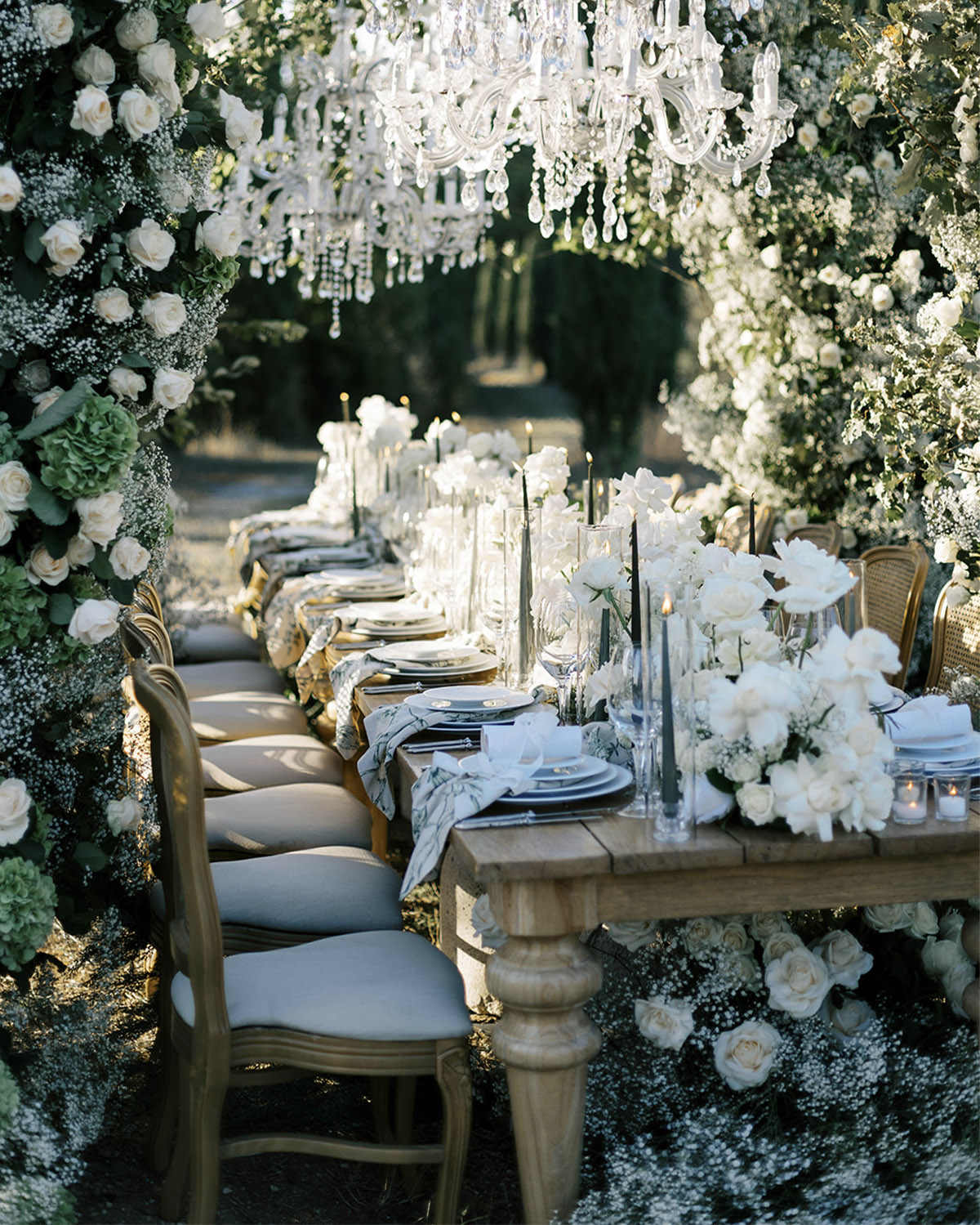 A romantic outdoor reception at an Italian villa garden features a long wooden table draped in white florals and glassware beneath cascading crystal chandeliers, styled in an elegant editorial wedding aesthetic.