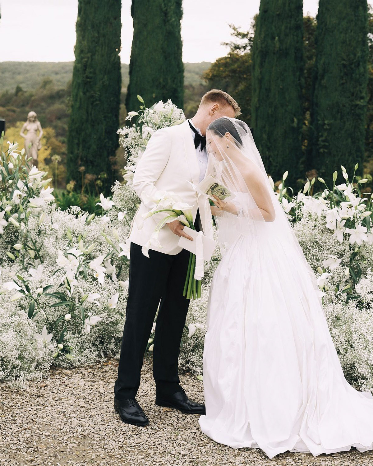 In a Tuscan countryside garden, a bride in a flowing veil and a groom in a white dinner jacket lean toward each other while holding white lilies, captured in a soft, editorial-style wedding portrait.