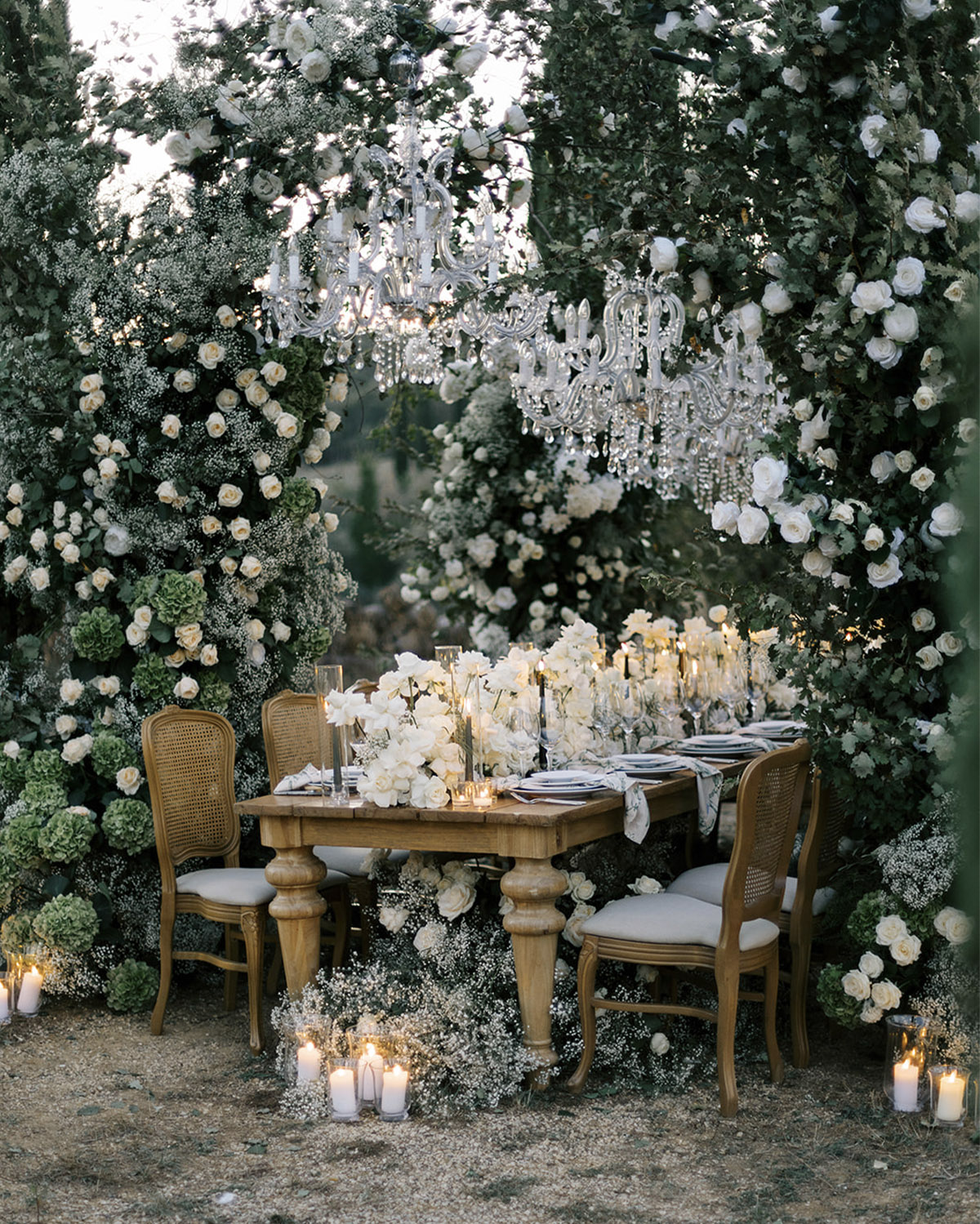 A lush garden setting at an Italian villa frames a long candlelit dining table surrounded by abundant white roses and greenery, with hanging chandeliers creating an editorial, fine-art wedding atmosphere.