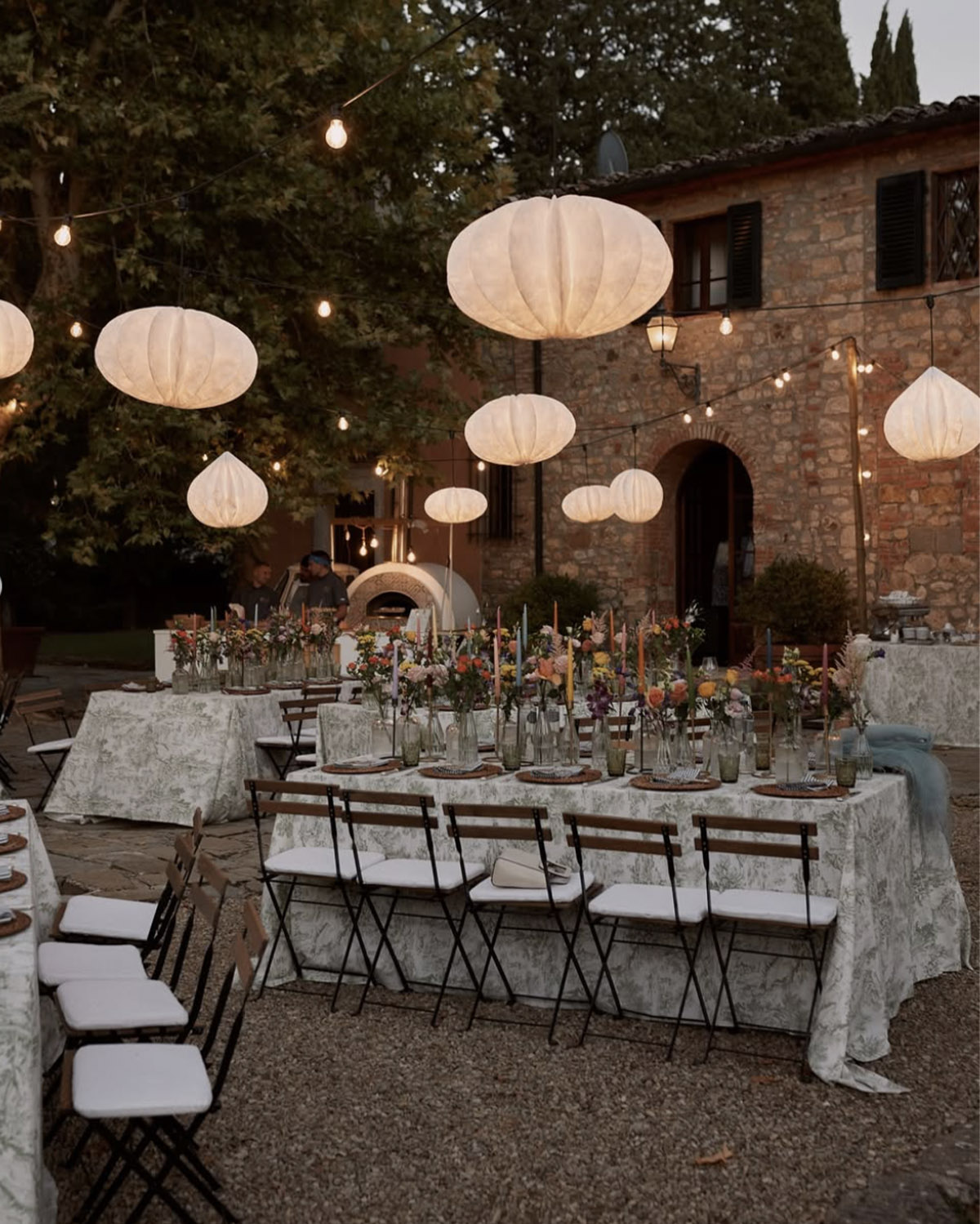 In a rustic Tuscan villa courtyard, long reception tables dressed in soft linens and colorful florals sit beneath glowing paper lanterns and string lights, captured in a warm, romantic editorial style.