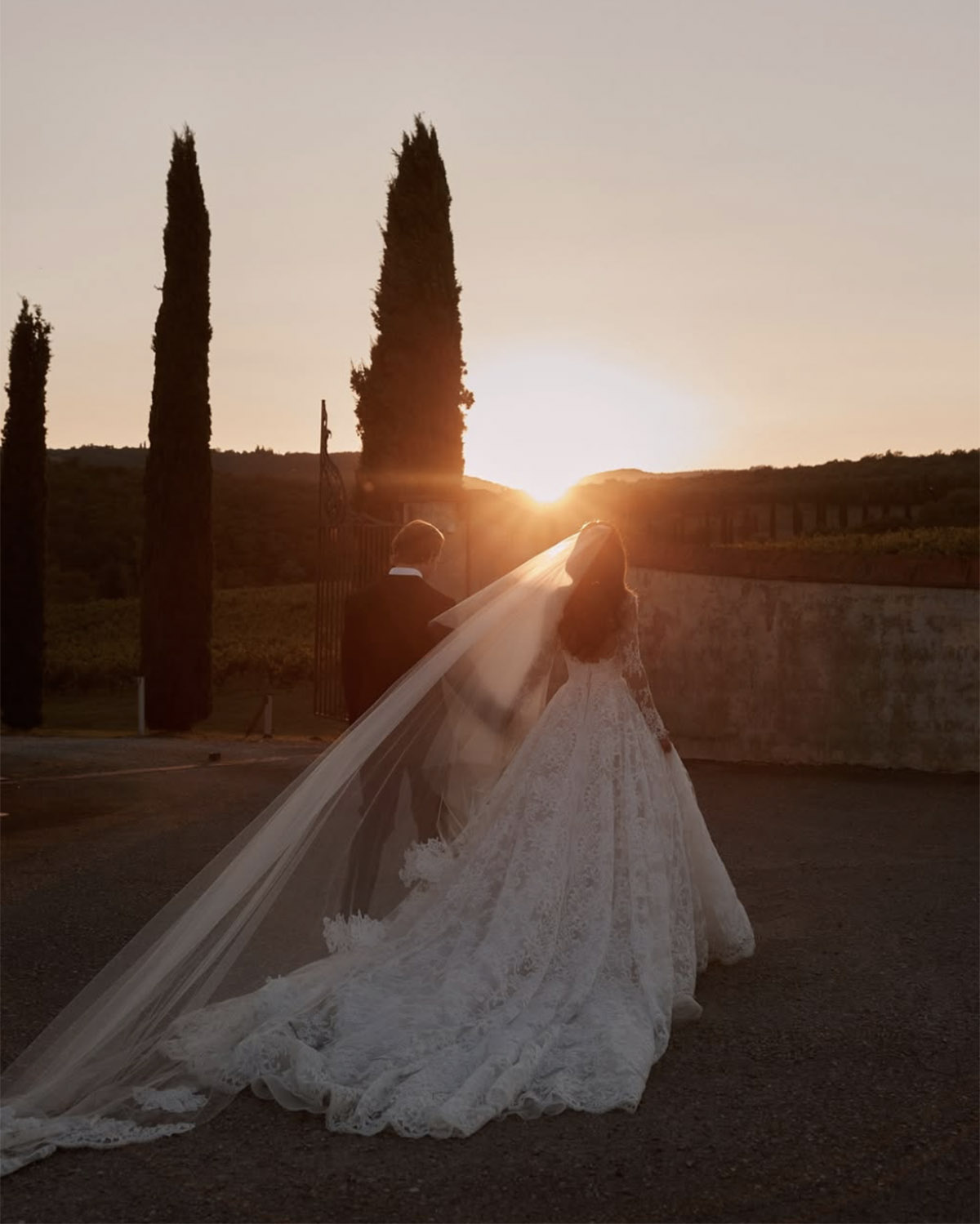Along a quiet countryside road in Tuscany at sunset, a bride in a flowing lace gown and long veil walks beside her partner toward the horizon, photographed in a cinematic, golden-hour editorial style.