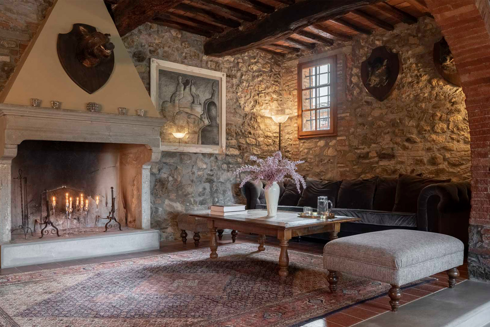 Inside a rustic Tuscan villa, a cozy stone-walled living room with a lit fireplace, wooden beams, and vintage furnishings is captured in a warm, editorial interior style.