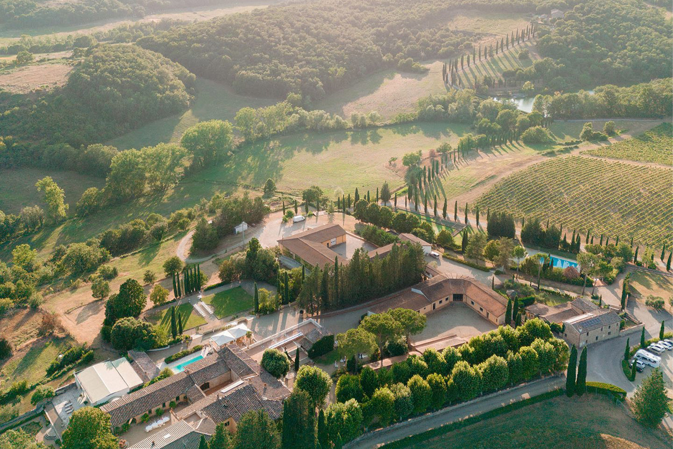 An aerial view over rolling hills in Tuscany, Italy, reveals a countryside estate surrounded by vineyards, cypress-lined roads, and golden fields in a dreamy, editorial landscape style.