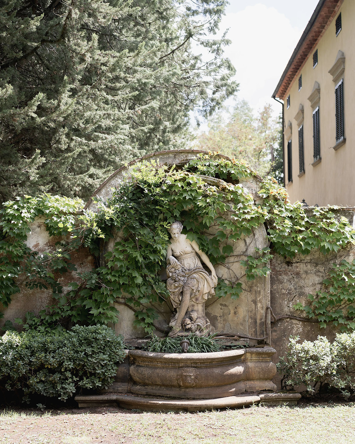 A quiet Tuscan villa garden showcases a weathered stone fountain with a classical statue, wrapped in ivy and surrounded by greenery, styled like a timeless editorial landscape photograph.