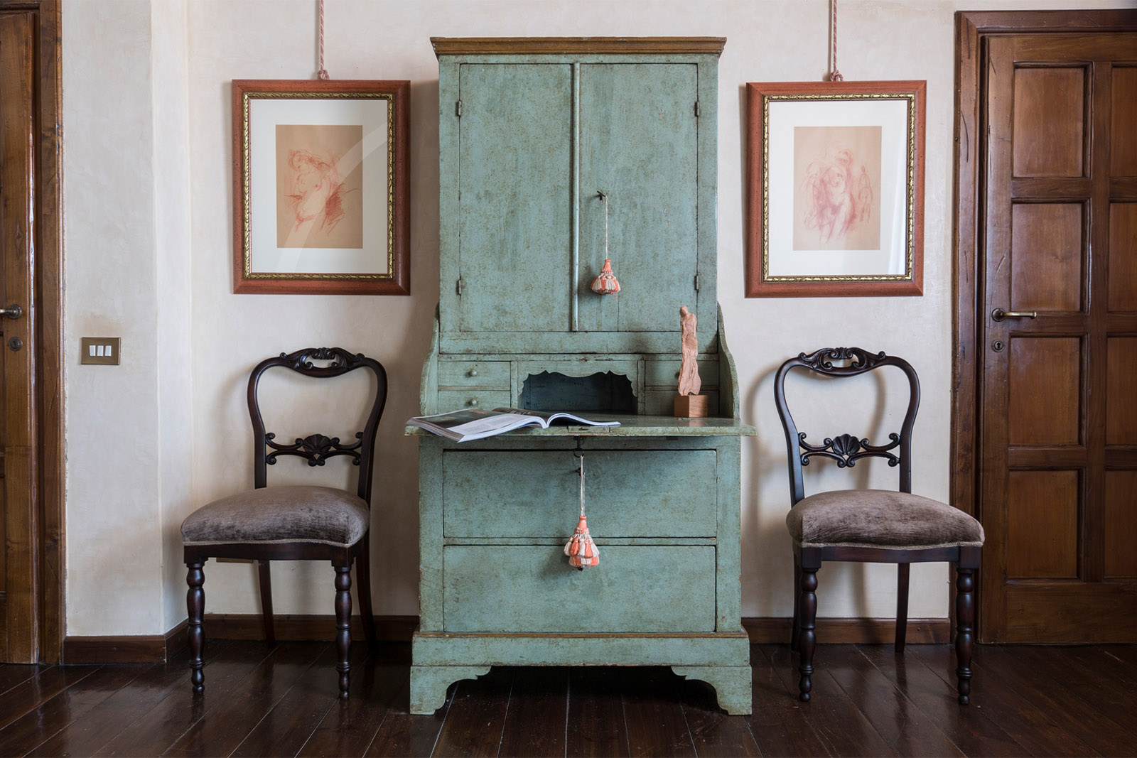 Inside a Tuscan villa, a symmetrical vignette of a vintage green writing cabinet flanked by two chairs and framed sketches is presented in a refined, editorial interior style.