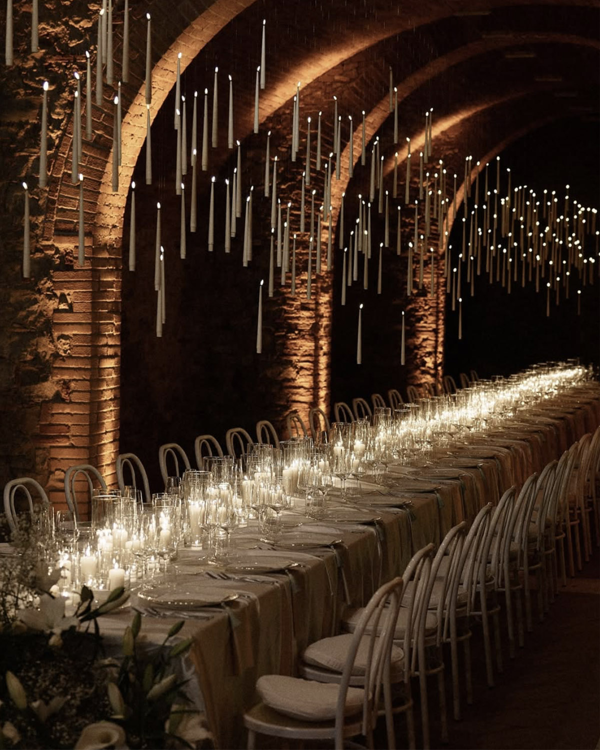 Inside a historic stone wine cellar in Tuscany, an elegantly set banquet table glows with candlelight beneath a ceiling of suspended taper candles, styled in a moody, atmospheric editorial aesthetic.